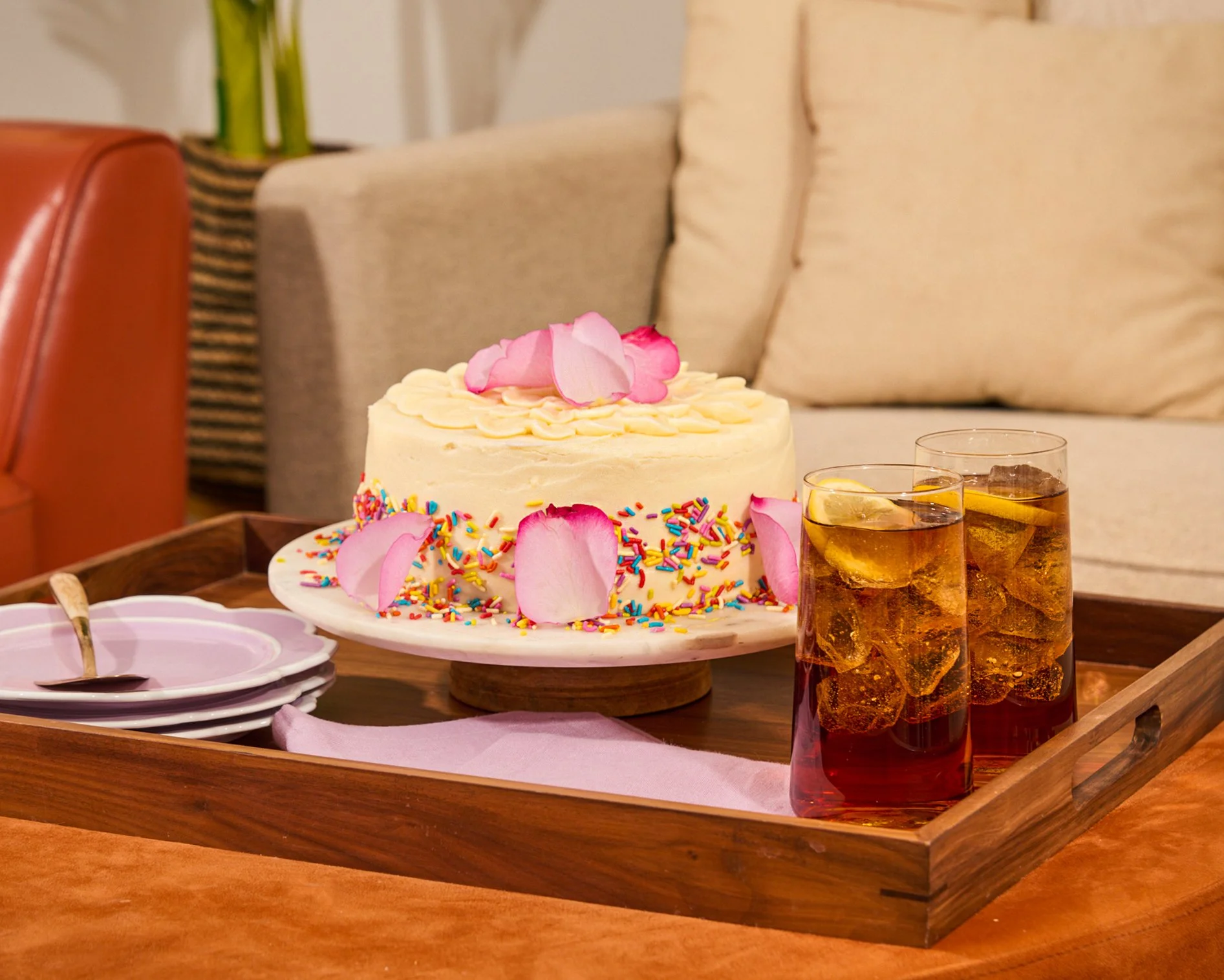 A birthday cake decorated with pink rose petals and colorful sprinkles on a wooden tray, with two glasses of iced tea, a stack of small plates, and a pink napkin, set on a wooden table in a cozy living room.