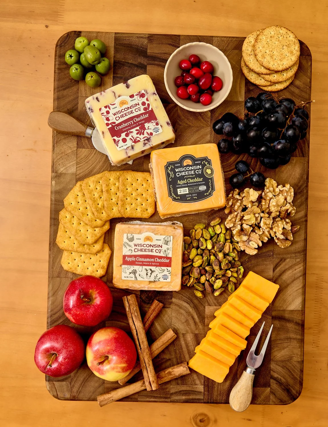 Wooden cheese board with green grapes, cranberries, three kinds of cheese, crackers, black grapes, walnuts, pistachios, apple slices, apples, cinnamon sticks, and cheese fork.