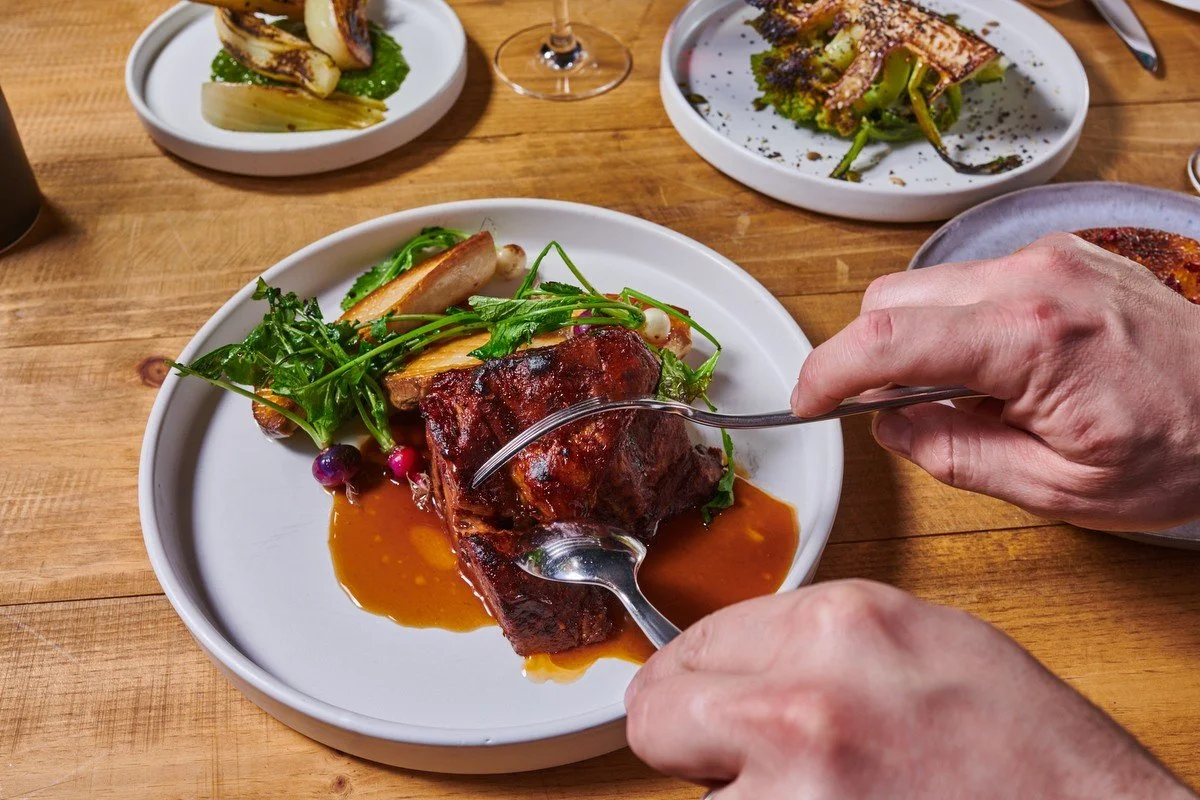 Hands cutting braised meat with sauce and greens, restaurant menu photography for the Culinary Institute of America