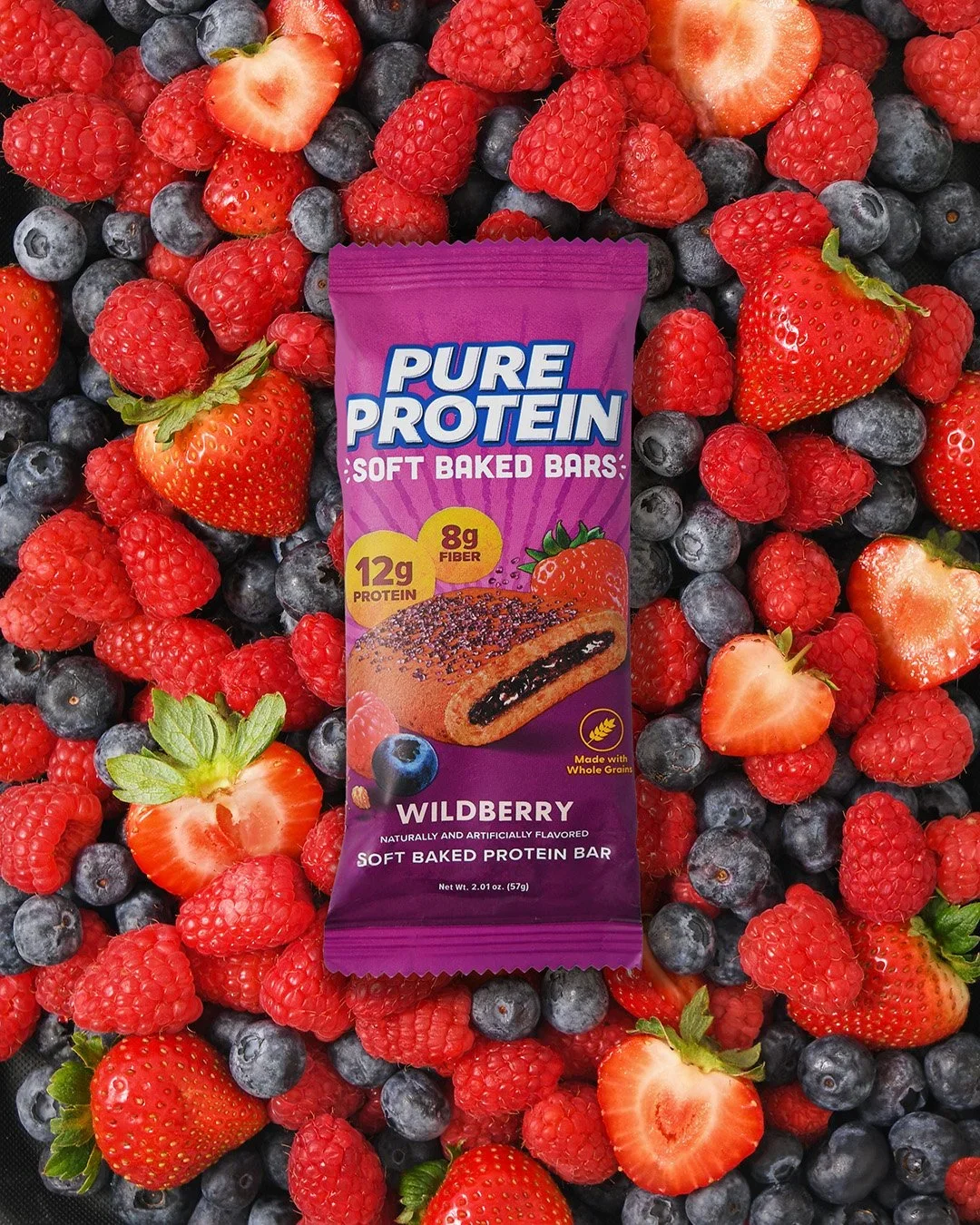A purple package of Pure Protein Wildberry soft baked protein bars placed on a bed of fresh strawberries, raspberries, and blueberries.