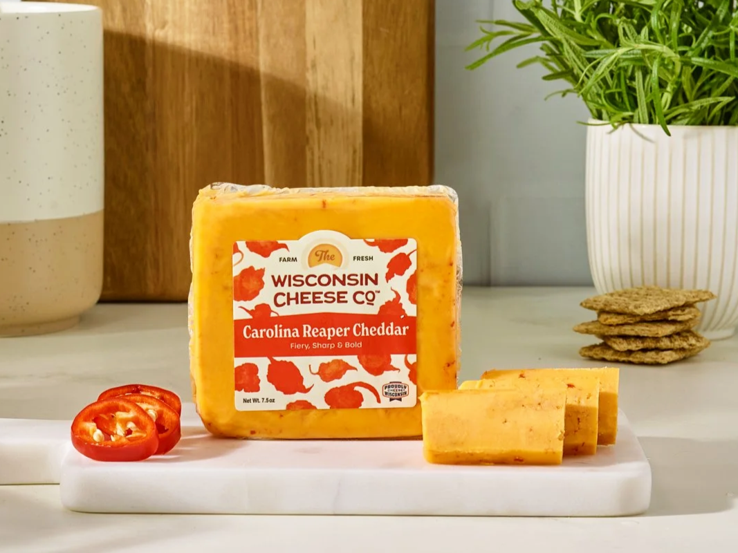 A block of Wisconsin cheese labeled 'Carolina Reaper Cheddar,' along with two slices of cheese, a slice of tomato, a potted plant, and some crackers on a white surface with a wooden background.