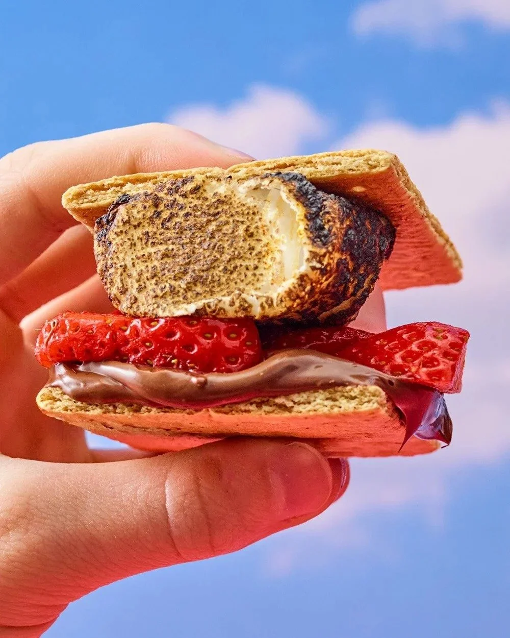 Hand holding a strawberry s'more with chocolate hazelnut spread against blue sky, food photography