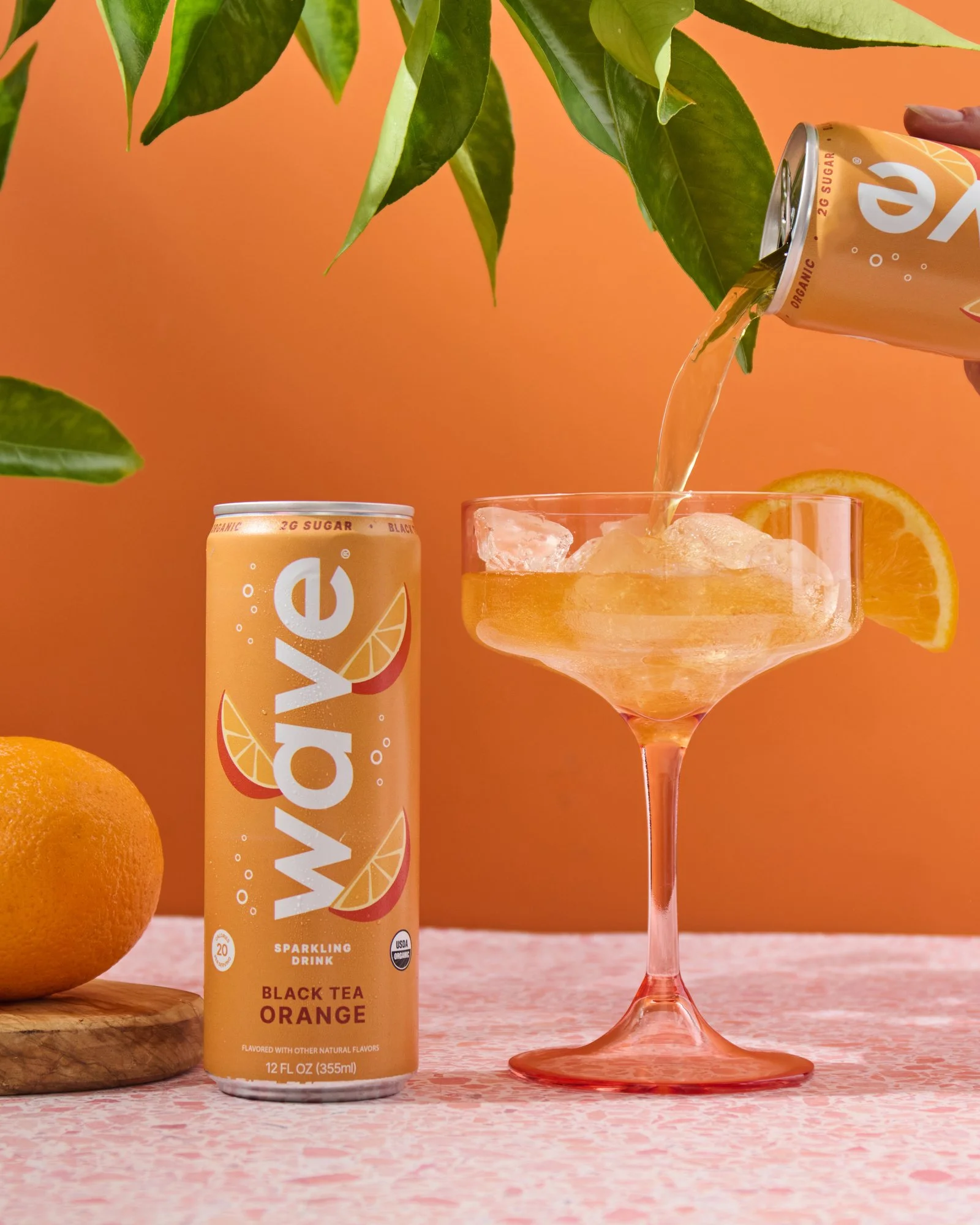 A can of Sparkling Orange Black Tea and a glass of orange-colored drink with ice, with a lemon wedge on the rim, on a pink patterned surface against an orange background, with a person pouring a can of sparkling orange soda into the glass and a whole