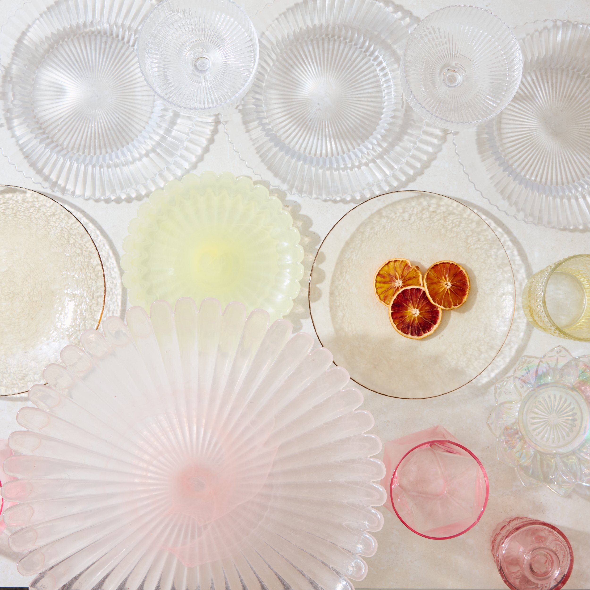 A collection of various glass dessert plates and bowls in clear, pink, yellow, and iridescent colors, with some dried citrus slices on one plate.