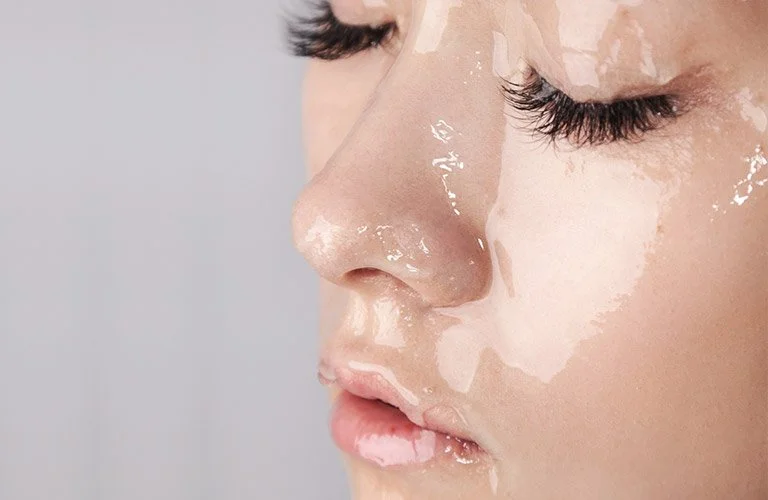 Close-up of a person's face with glossy skin and long eyelashes, showing a skincare or beauty treatment in progress.