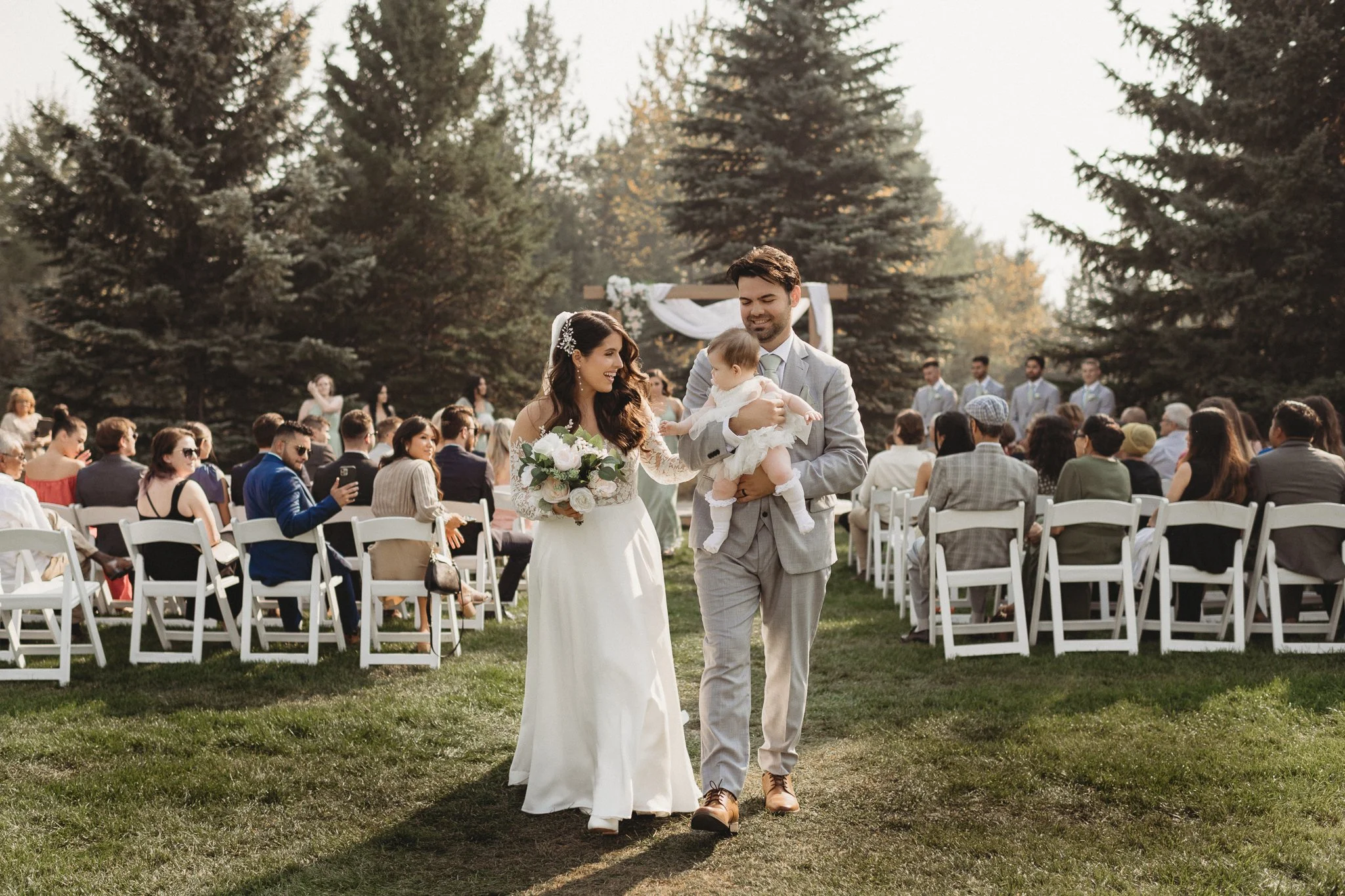 A couple walking down the aisle with a baby girl during an outdoor wedding ceremony, surrounded by seated guests and a backdrop of tall trees. Intimate wedding in Edmonton captured by Afton Photography.