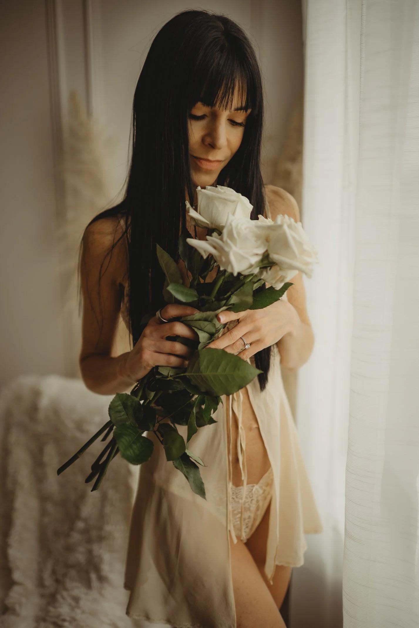 A woman with long dark hair and bangs holding white roses standing near a curtain, wearing beige lingerie and a silk robe.