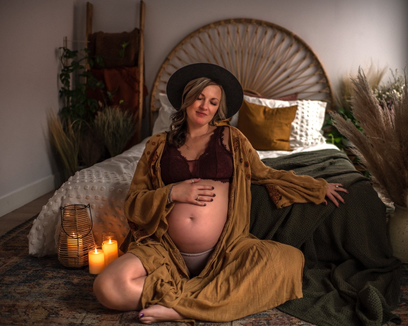 A pregnant woman with long wavy hair, wearing a black hat, lace top, and brown robe, sitting on the floor in a cozy room decorated with candles, plants, and pillows, with her eyes closed and a peaceful expression.