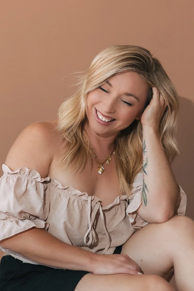 A smiling woman with blonde hair and light skin, wearing an off-the-shoulder beige top and black shorts, sitting against a plain background.