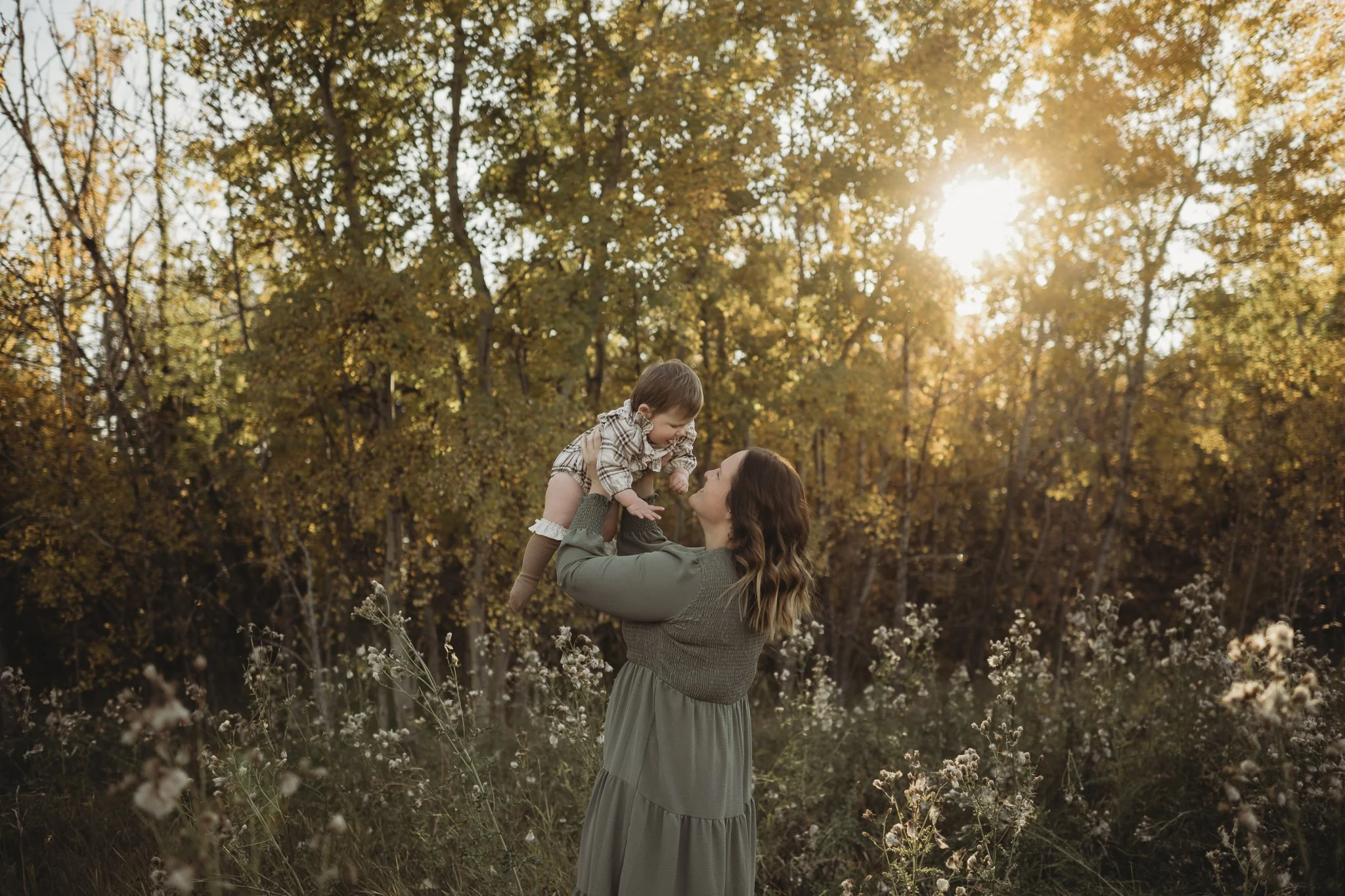 A woman lifting a child in a forest during sunset with golden sunlight filtering through trees.