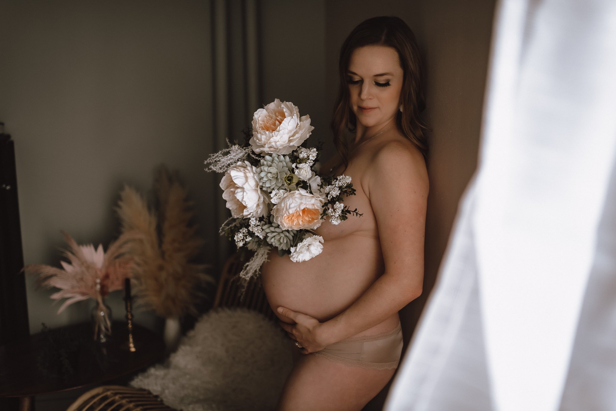 A woman with brown hair stands naked in front of a mirror, holding a large bouquet of white and light pink flowers. She is in a softly lit room with decorative dried plants in the background.