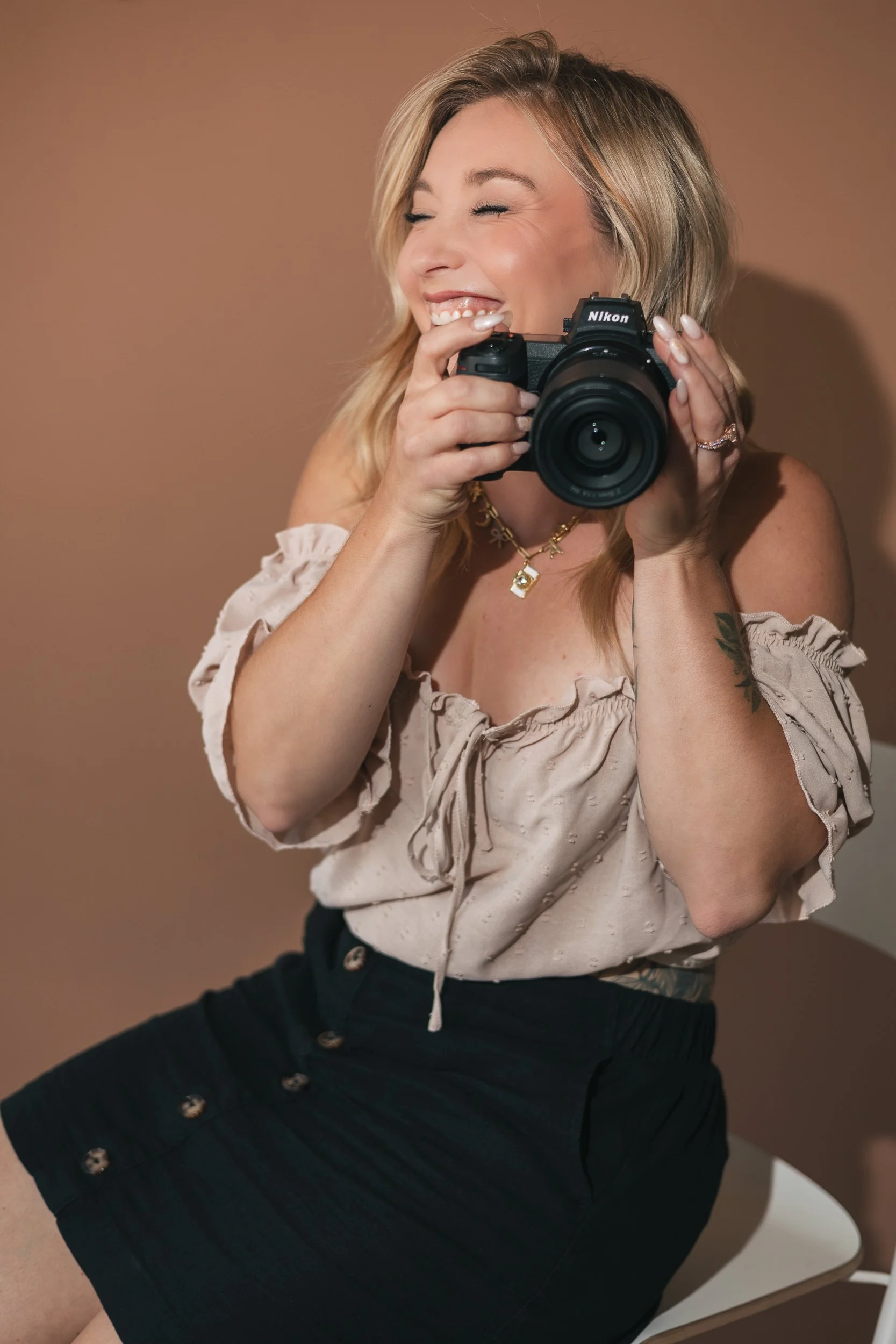 A woman with blonde hair taking a photo with a Nikon camera, wearing a beige off-the-shoulder blouse and a black skirt with buttons.
