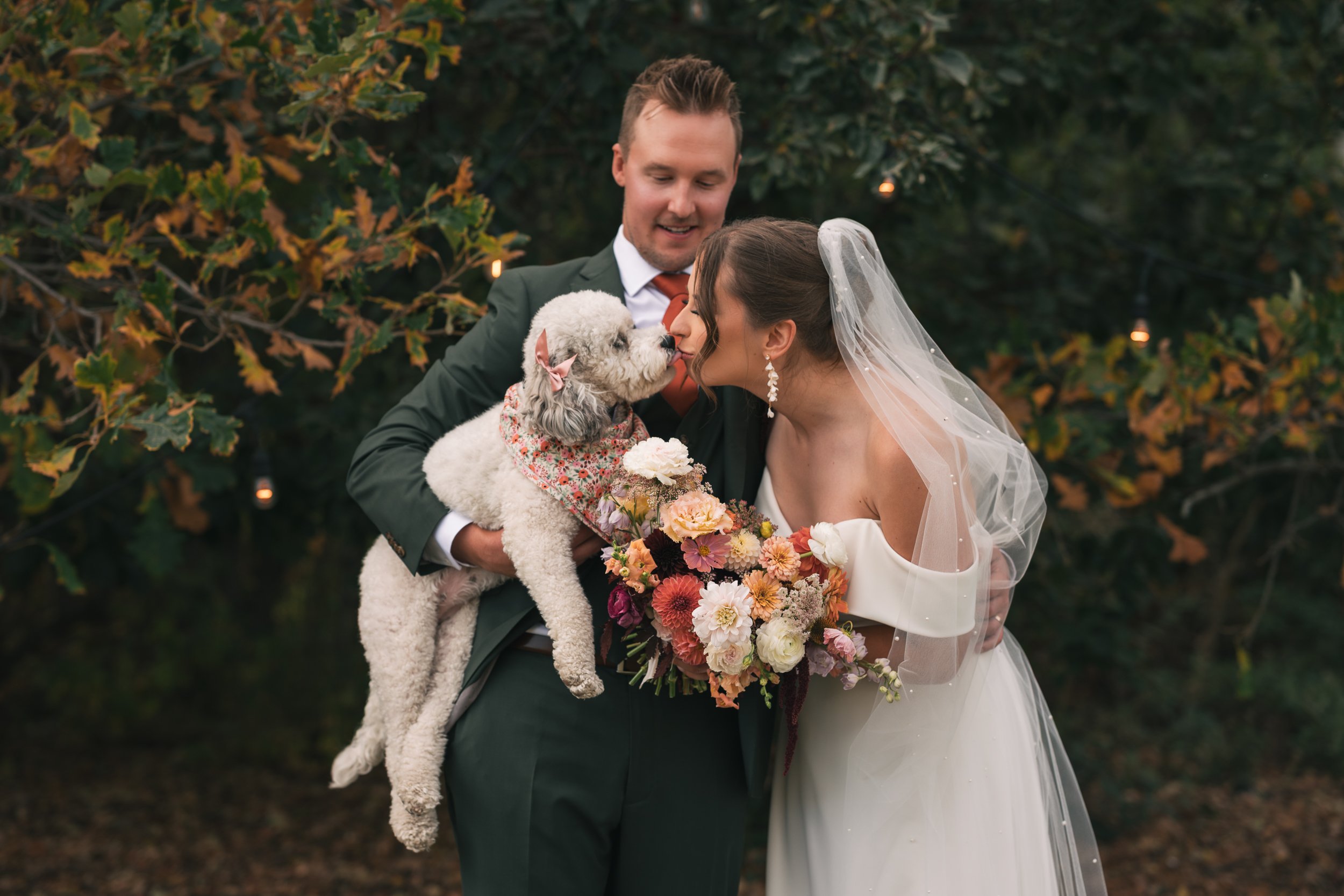 A bride and groom holding a dog during a wedding ceremony in an outdoor garden setting, surrounded by greenery and string lights.