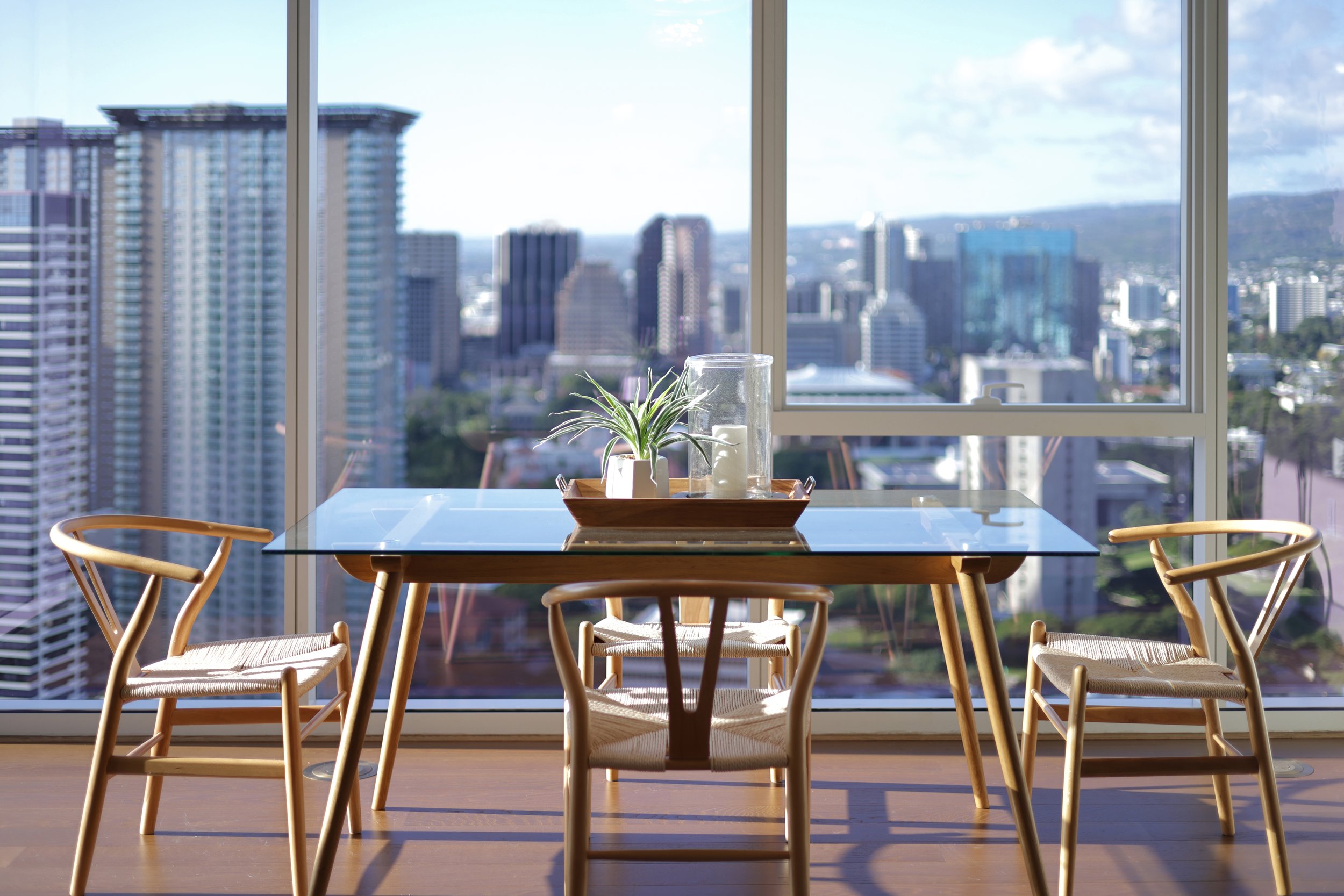 floor to ceiling glass windows overlook downtown honolulu. an all wood set of dining chairs surrounds a glass topped dining table with a service tray in the center.