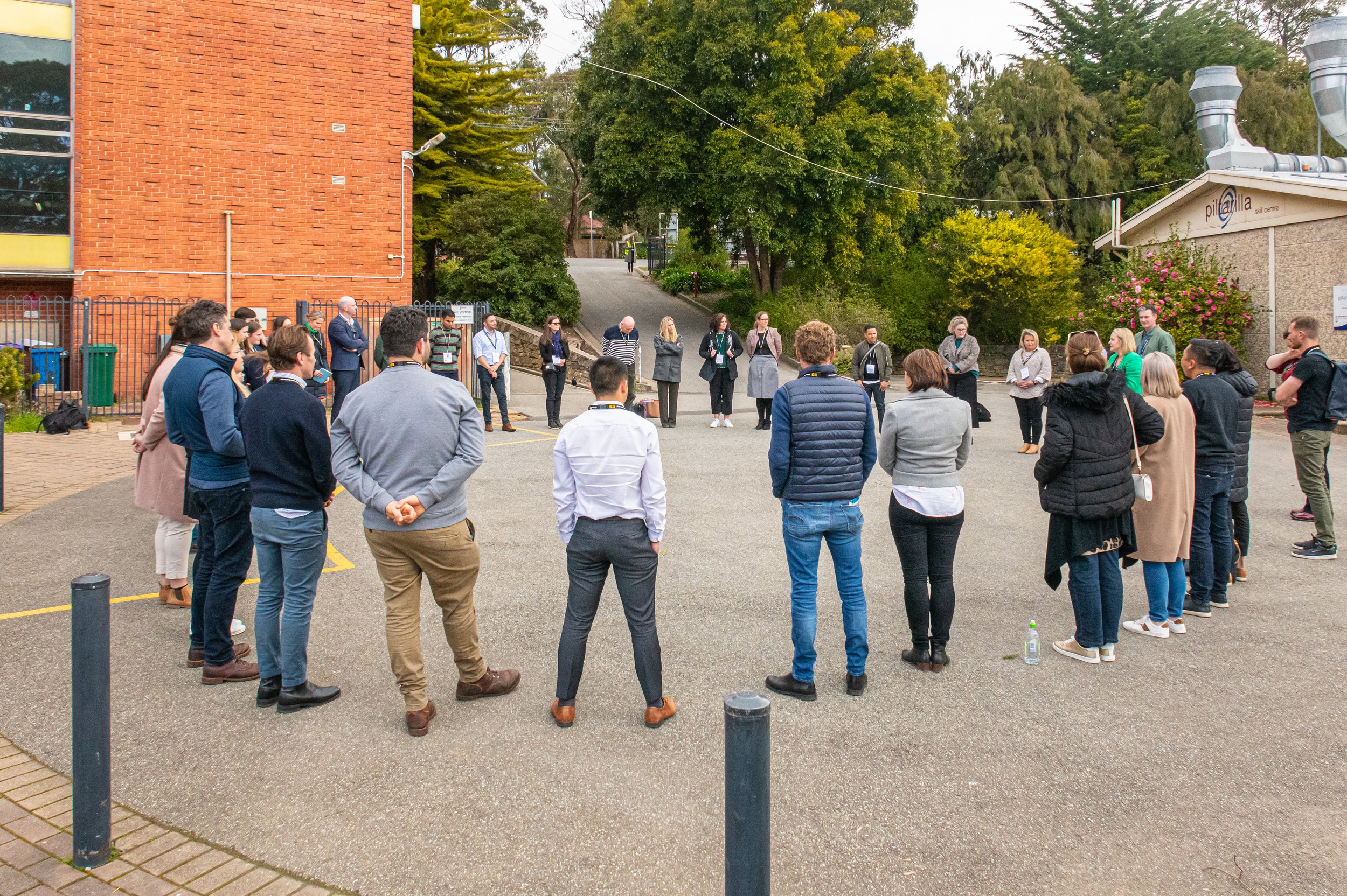 A group of people standing in a circle outside, participating in a discussion or meeting, with a building and trees in the background.