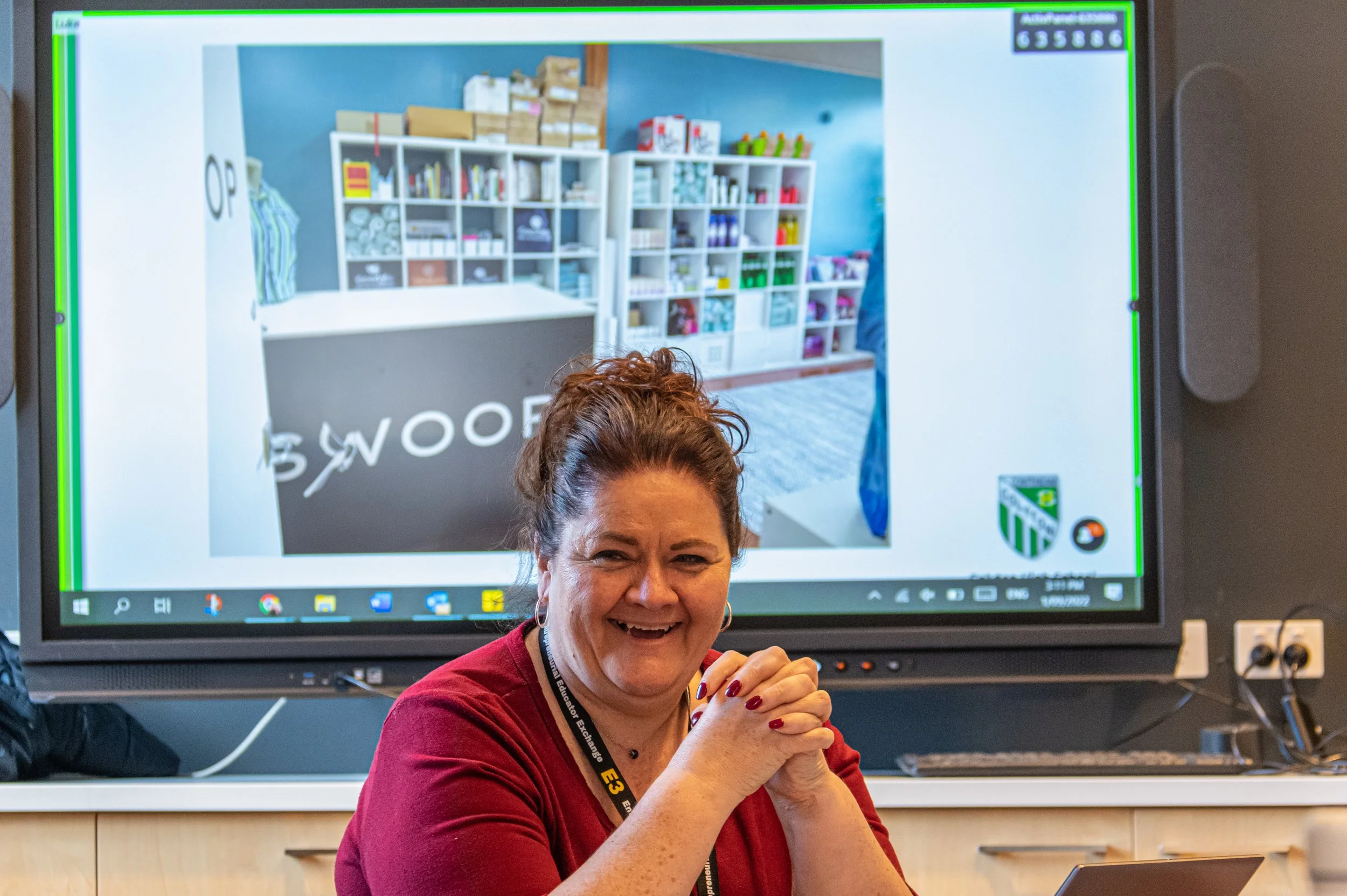 A woman with dark hair tied up, wearing a red shirt, smiling and sitting at a desk in a classroom. Behind her, a large computer monitor displays a classroom or office space with shelves filled with books, boxes, and various supplies.