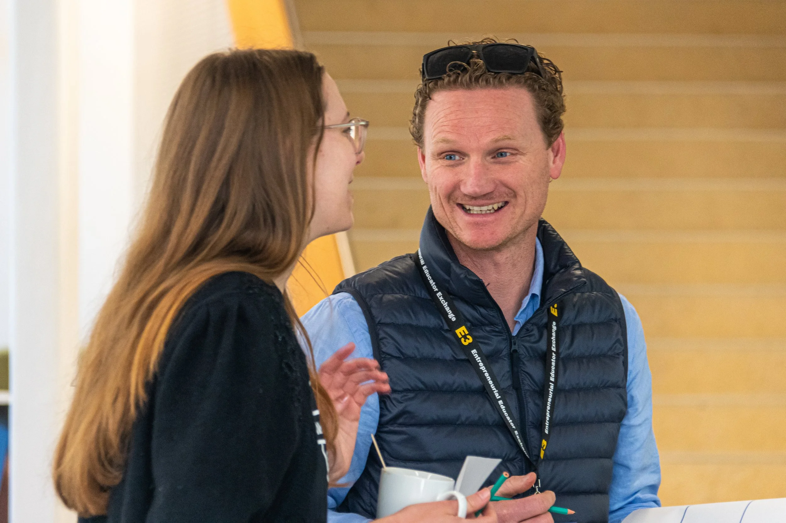A man and woman engaged in conversation indoors, smiling at each other. The man is wearing a blue shirt and a black insulated vest with a lanyard around his neck. The woman has long red hair and glasses, holding a white mug. The background shows wooden stairs.