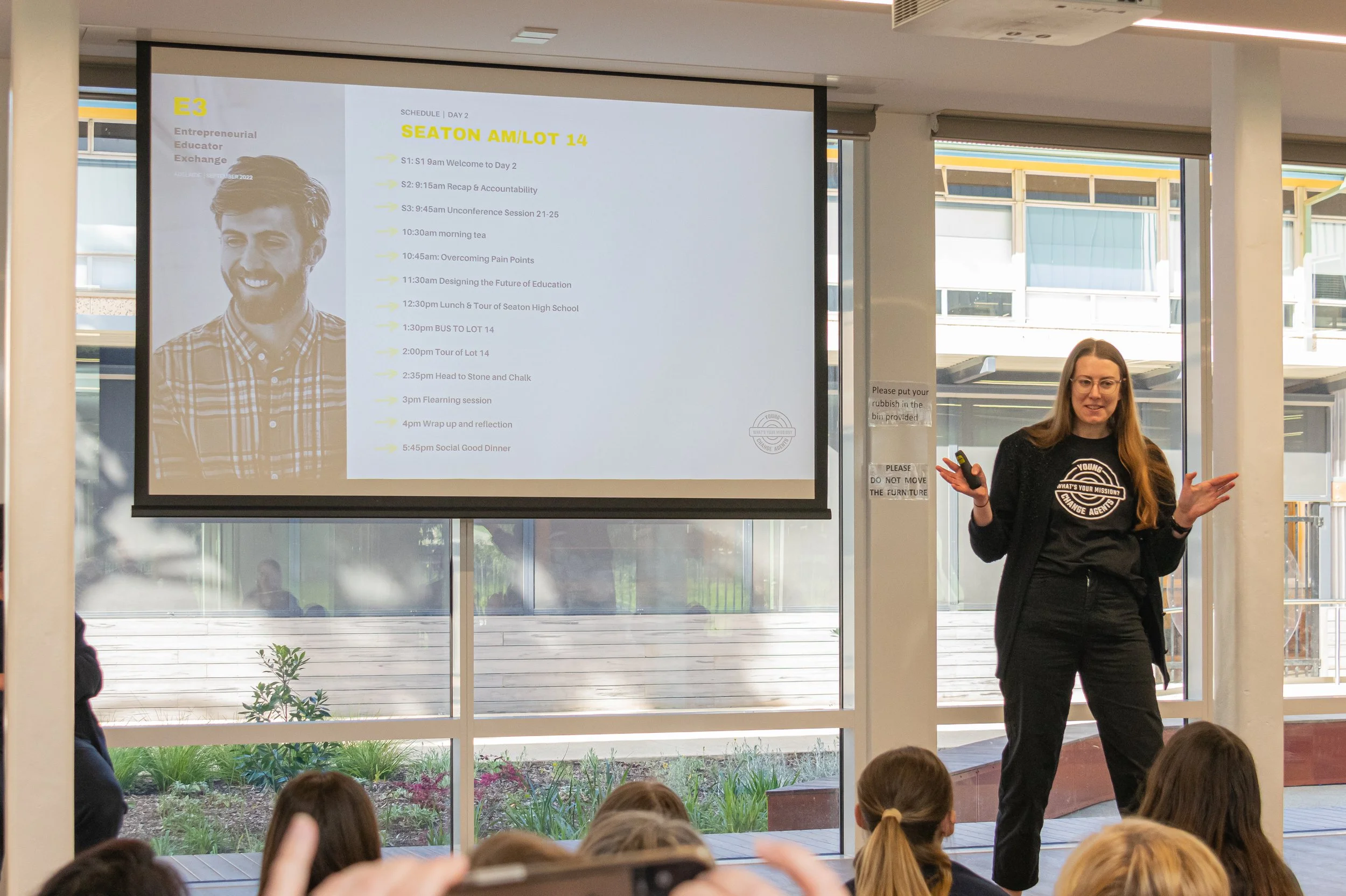 A woman giving a presentation to an audience with a slide projection behind her. The slide displays the schedule for a day at a conference, including times and activities. The slide also features a grayscale portrait of a smiling man.