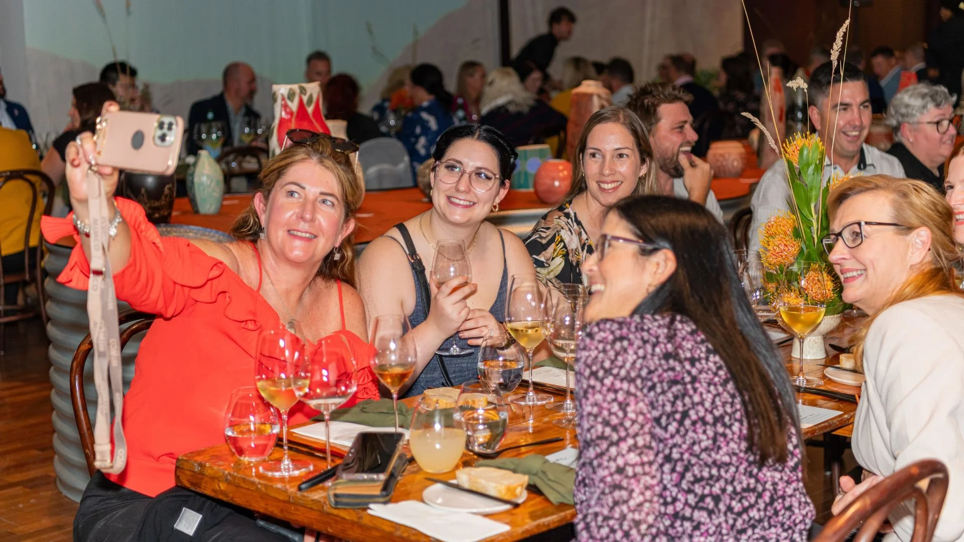 Group of smiling women taking a selfie at a table during a social event. They have glasses of wine and enjoyed food, with floral arrangements and other guests in the background.