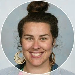 A woman with dark hair styled in a bun, smiling, wearing large gold earrings and a dark top, against a light blue background.
