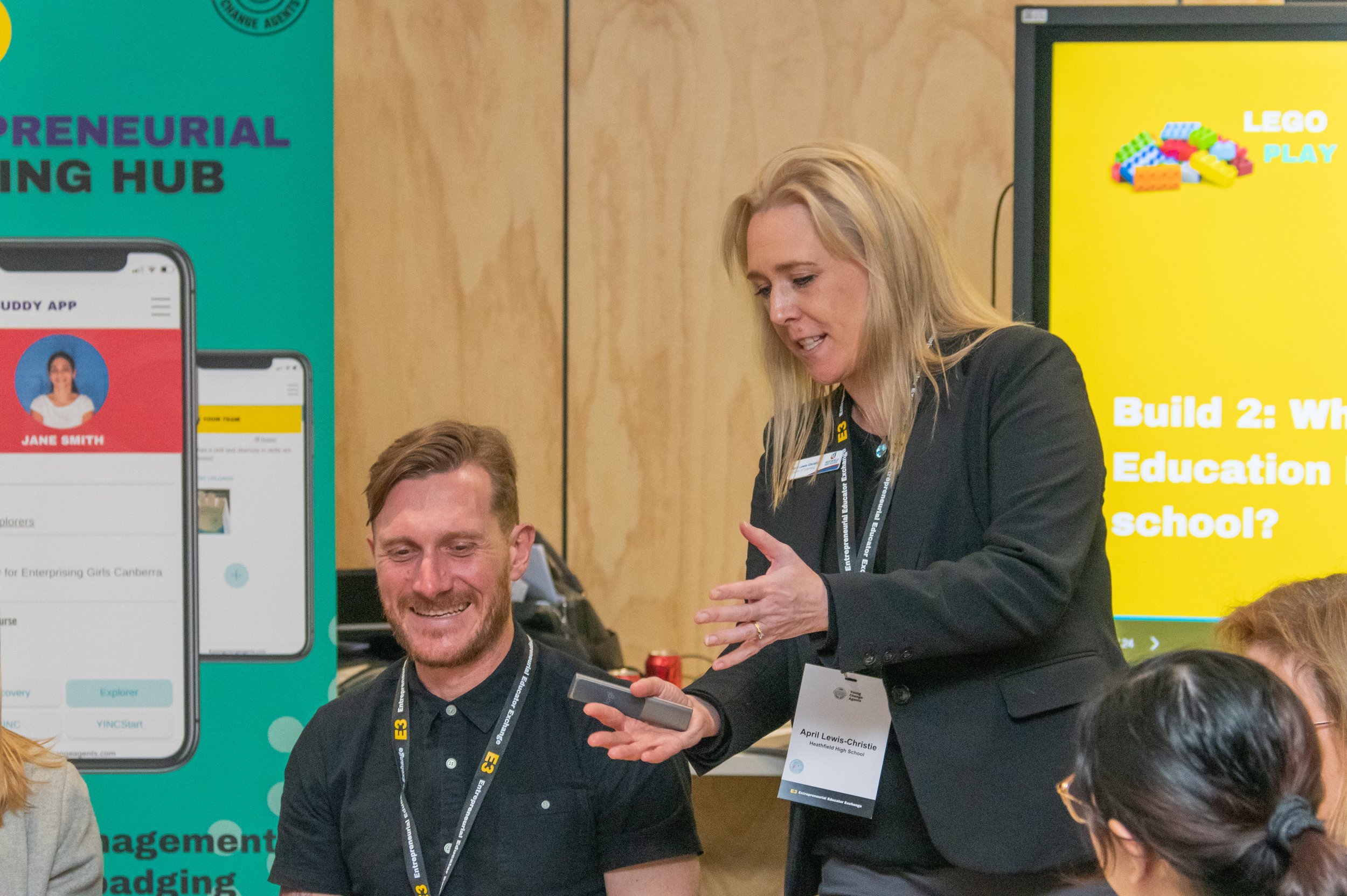A woman with blonde hair wearing a black blazer shows something on her phone to a man with light brown hair and a beard, who is smiling. They are at a professional event with banners and a large yellow screen in the background.