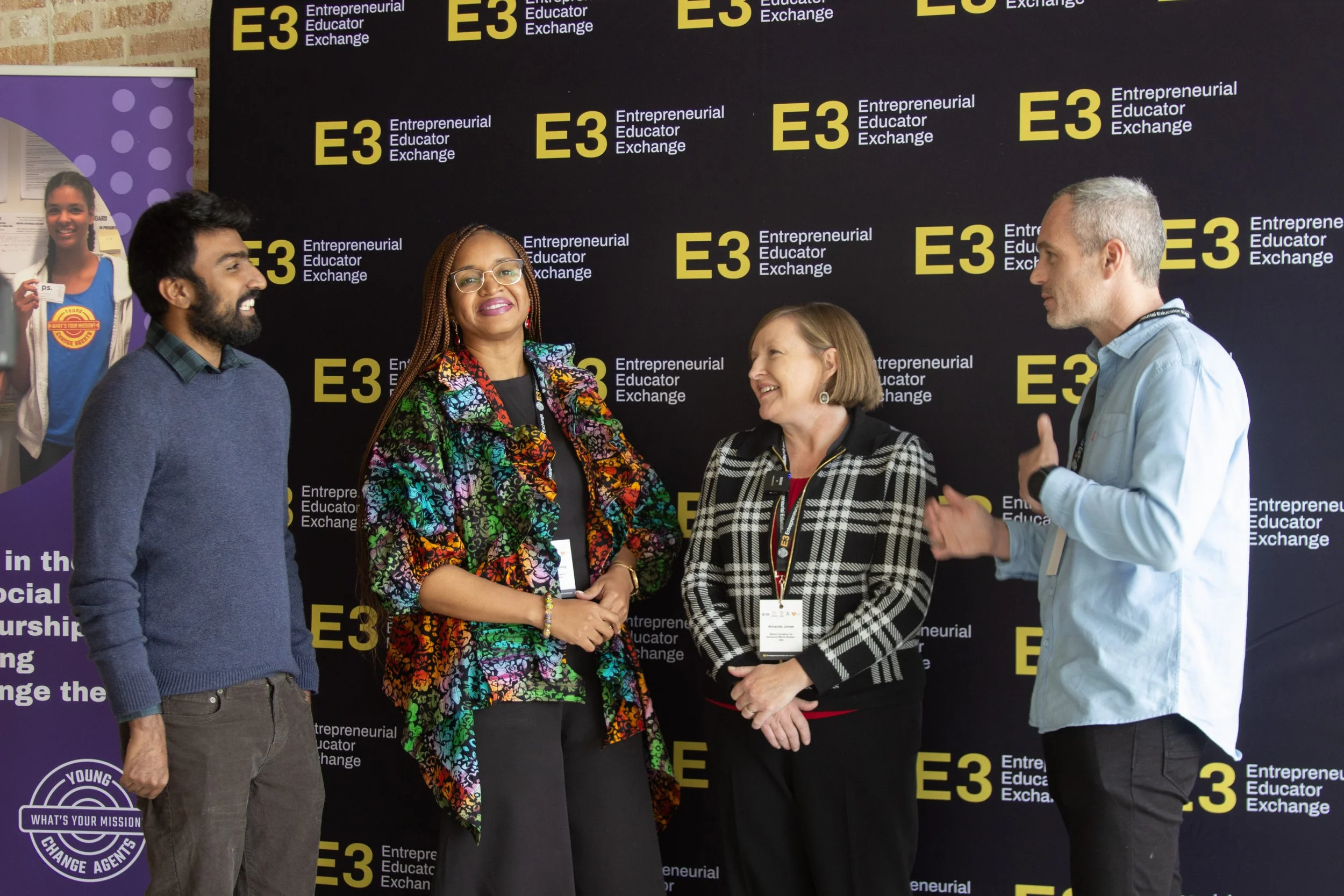 Four diverse people standing and talking in front of a black backdrop with yellow lettering that reads "E3 Entrepreneurial Educator Exchange." One woman is wearing a colorful patterned jacket, another a black and white checkered blazer, a man in a light blue shirt, and a man in a blue sweater. They are smiling and engaging in conversation.