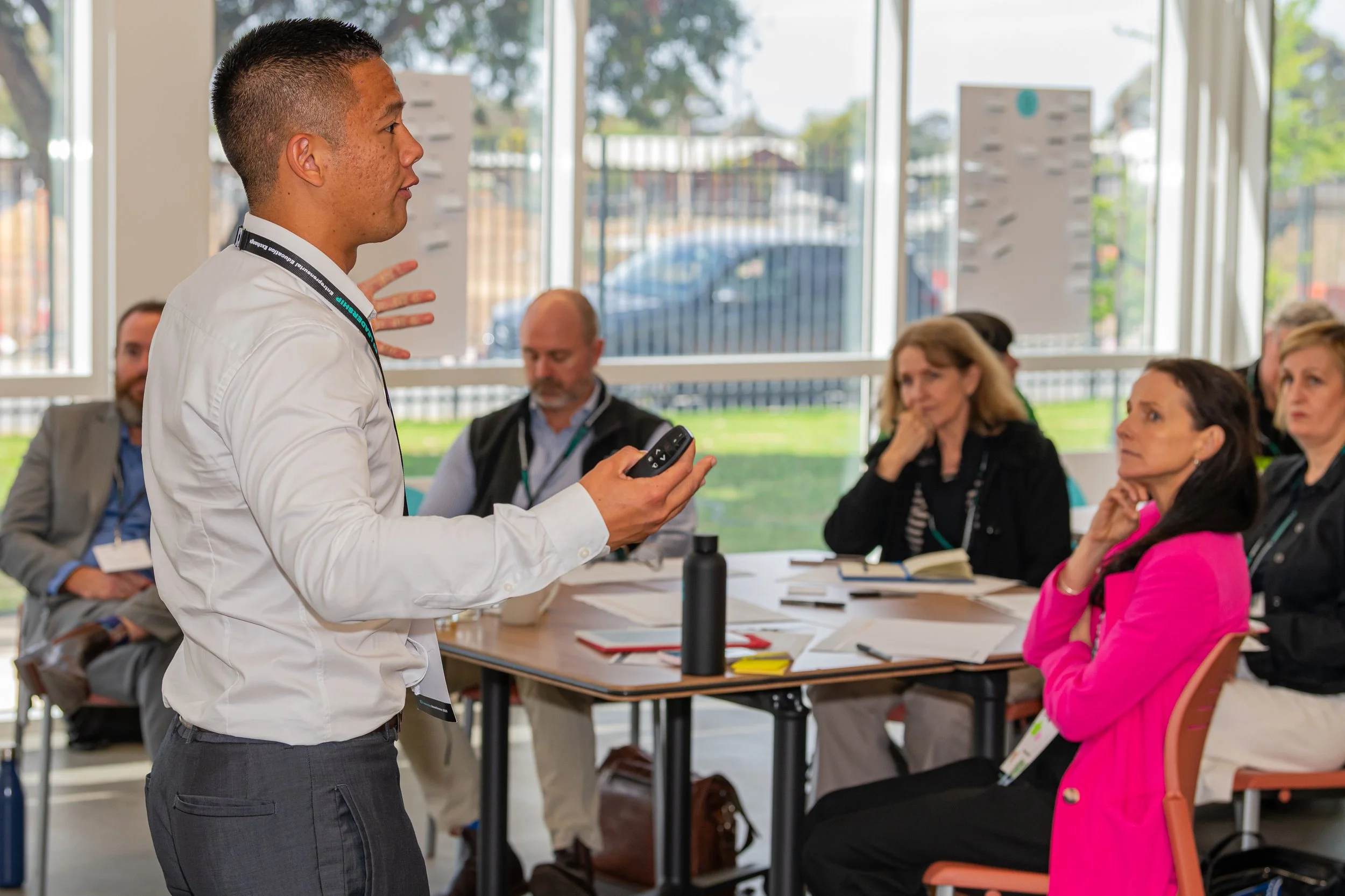 A young man in a white shirt is standing and addressing a group of people seated at a table during a meeting or presentation in a brightly lit room with large windows. The seated individuals are listening attentively, with some taking notes.