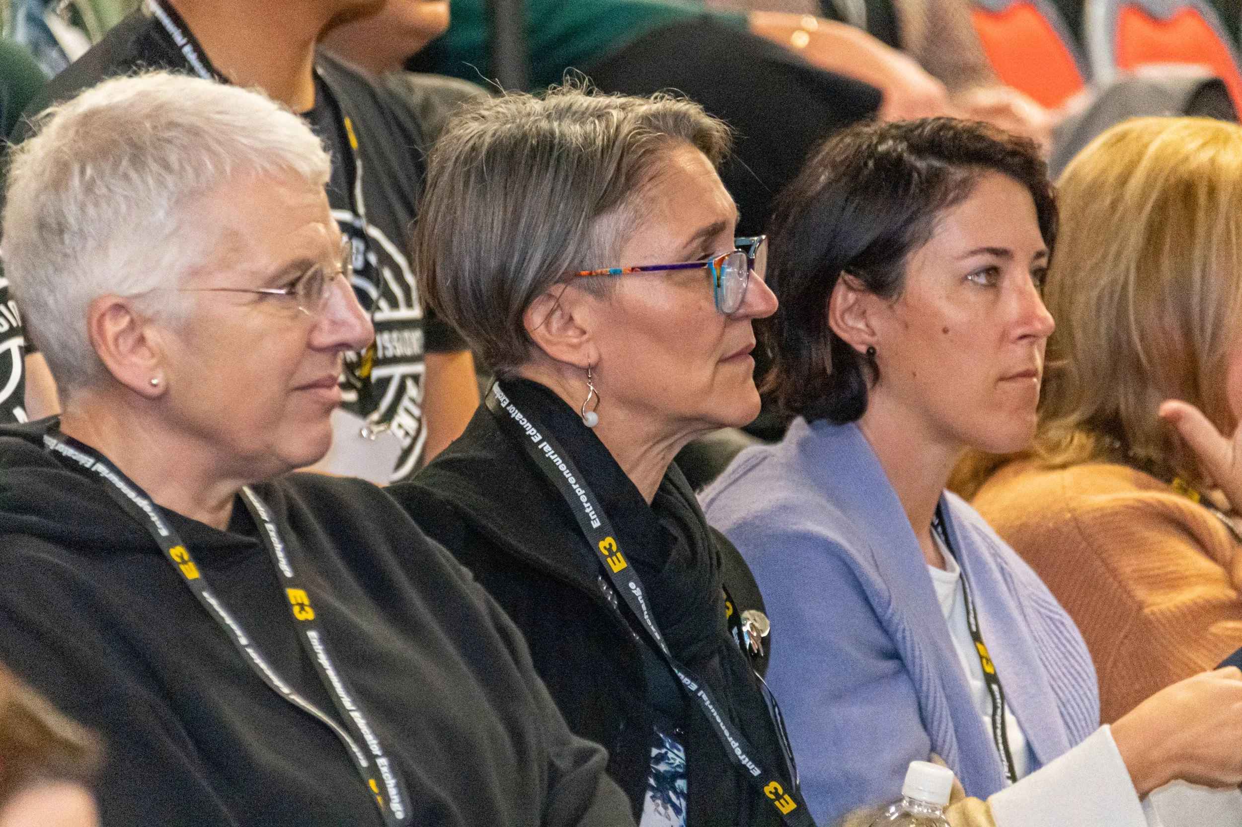 Three women seated at a conference, wearing conference badges with 'E3' on lanyards, listening attentively.