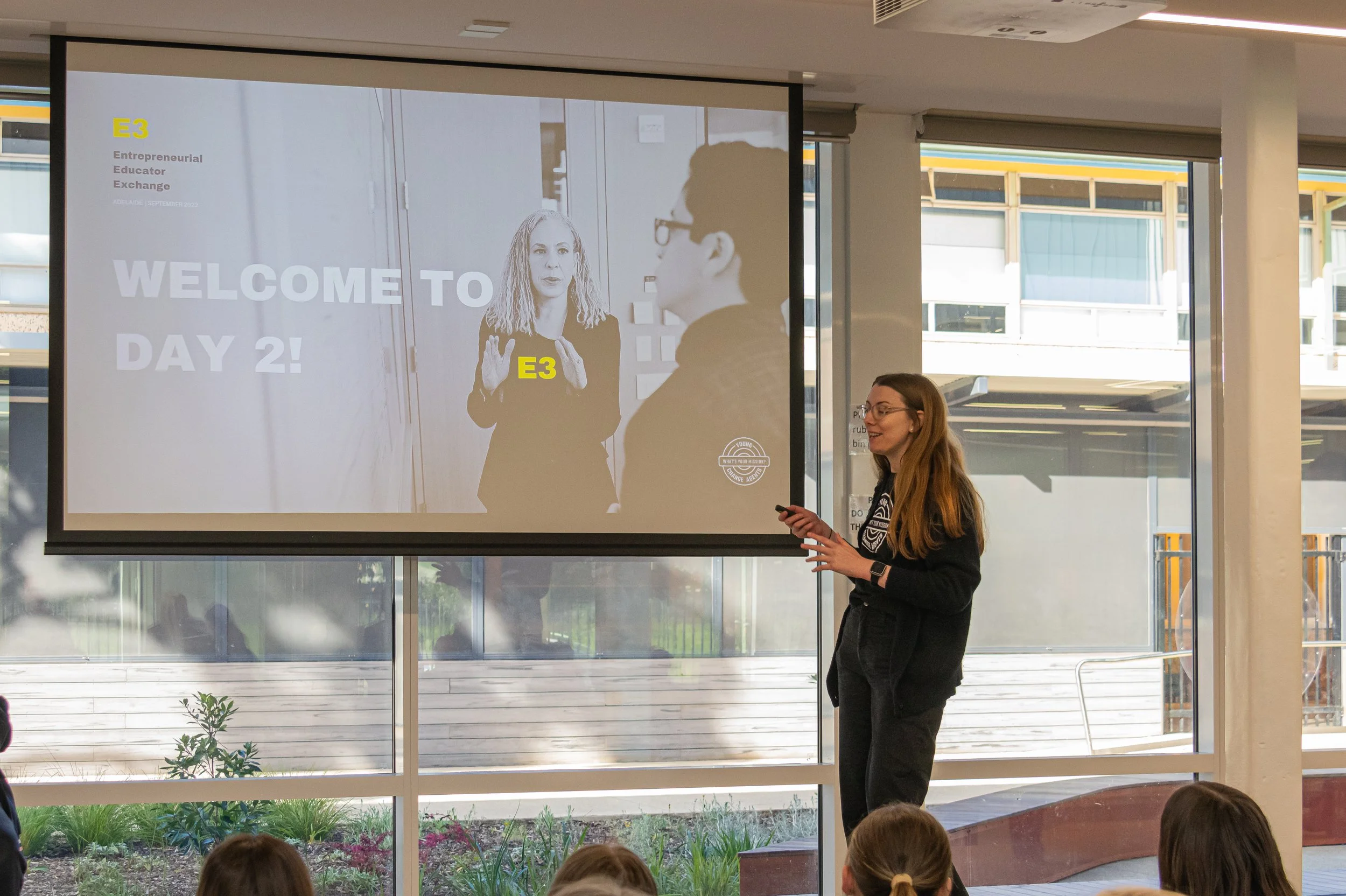 A woman is giving a presentation in front of a screen, with children seated in front listening. The screen shows a slide with the text 'WELCOME TO DAY 2!' and 'E3' in yellow, along with images of two people talking.