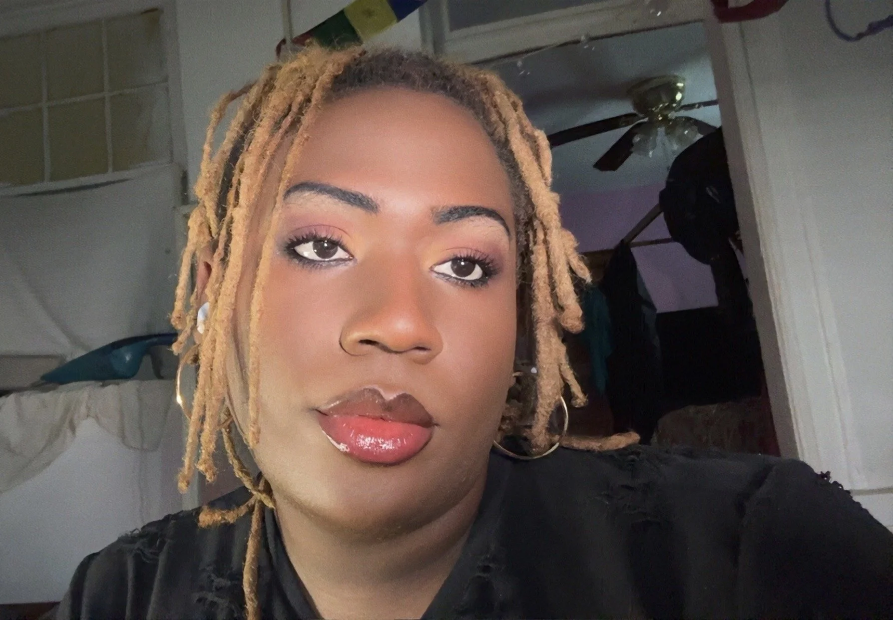 A woman with reddish-brown dreadlocks, wearing purple eyeshadow, red lipstick, and gold hoop earrings, smiling at the camera.