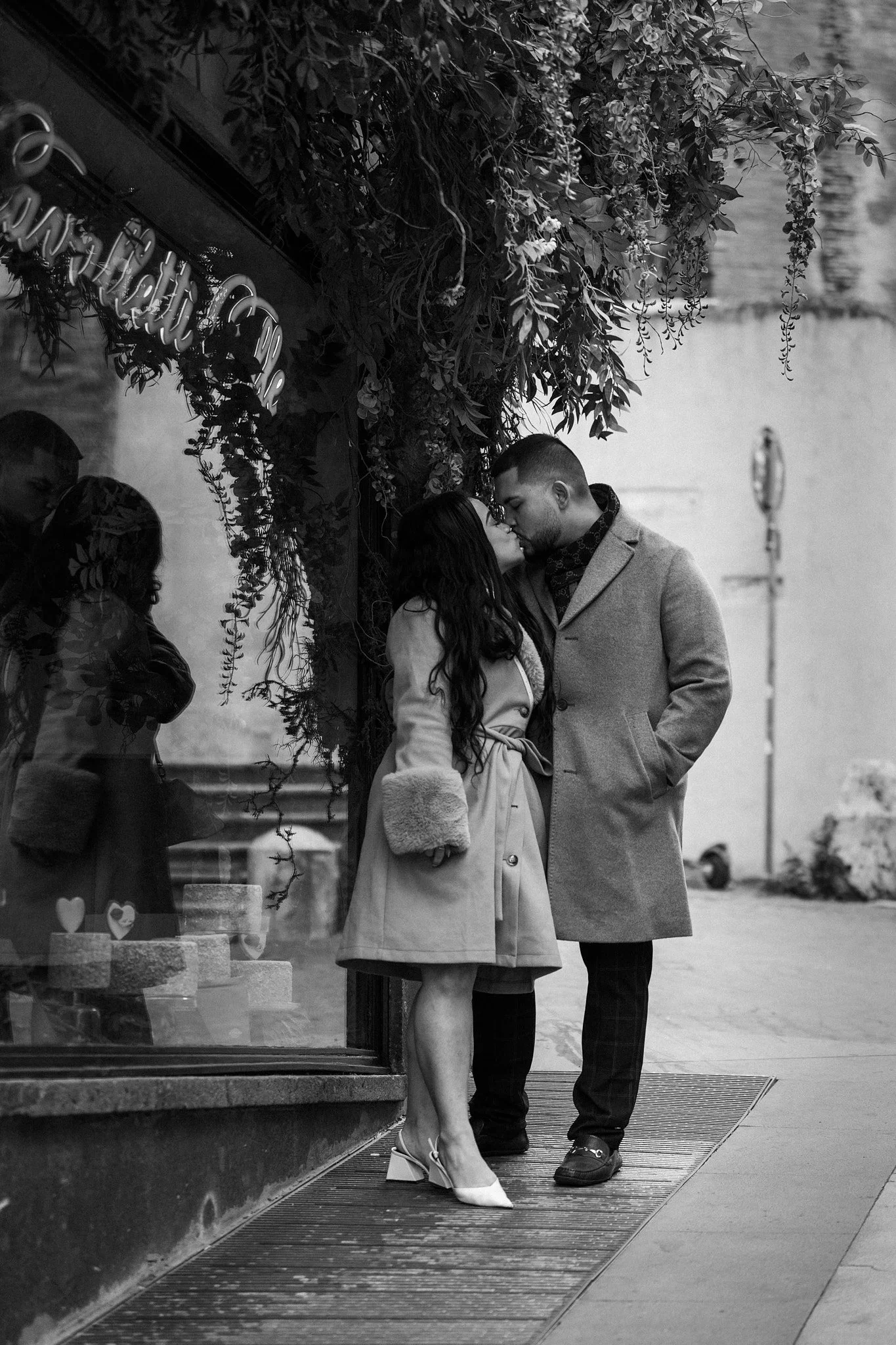 A couple is kissing on a city sidewalk in front of a store window decorated with greenery. The woman is wearing white high heels and a light coat, while the man is dressed in dark shoes, dashed pants, and a light overcoat. Other people can be seen re