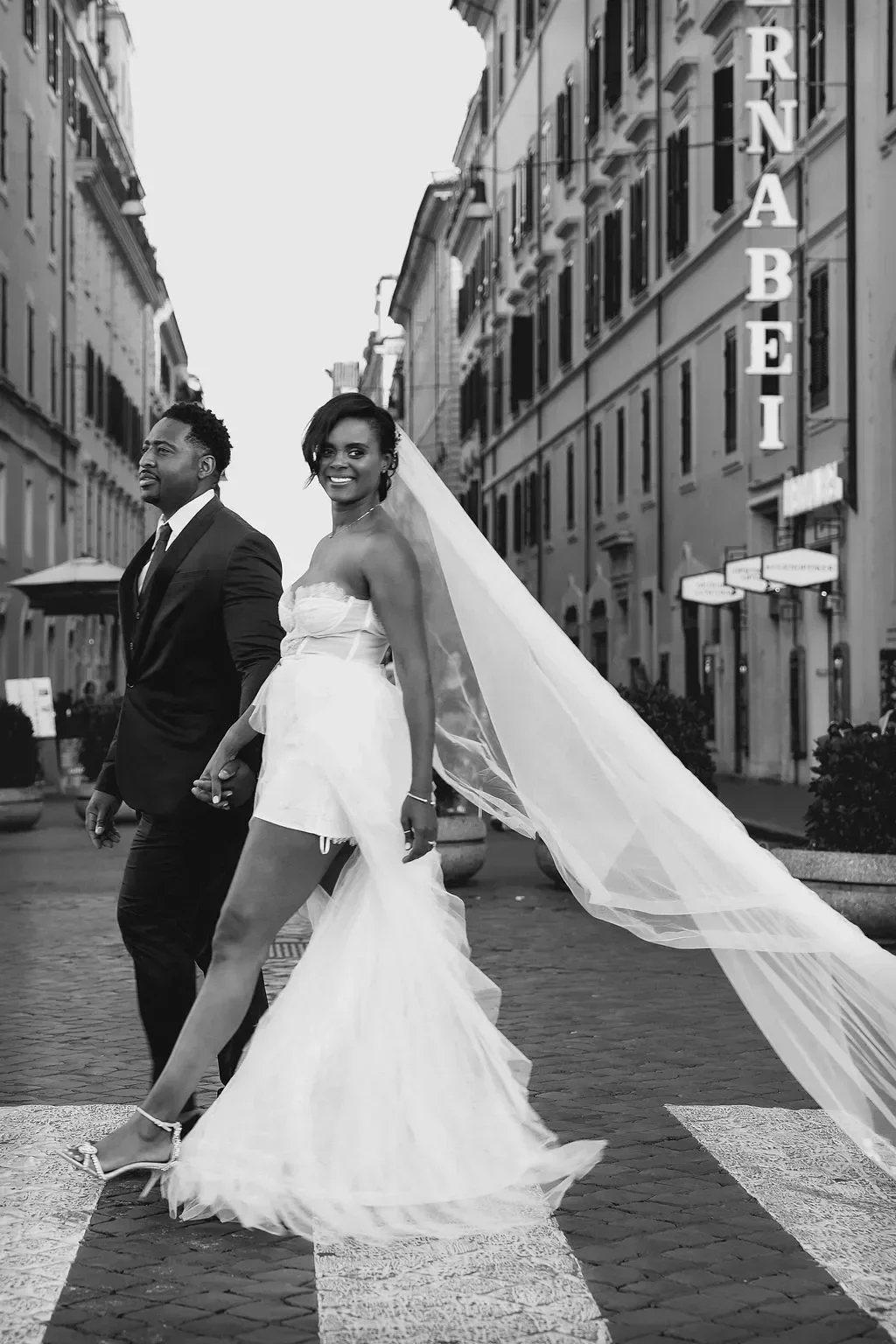 A bride and groom holding hands walking across a city street in black and white, with the bride smiling and wearing a wedding dress and veil, and the groom in a suit. The street has tall buildings and a sign that says 'RABEI' in the background.