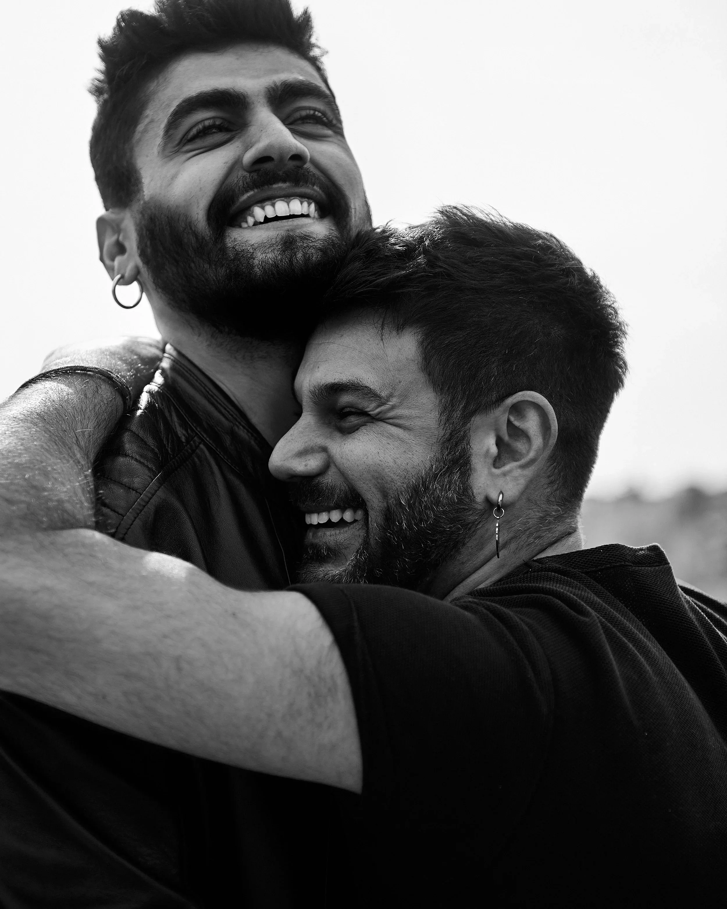 Two smiling young men hugging each other, one with a beard and earrings, both with dark hair, in a close-up black and white photo.