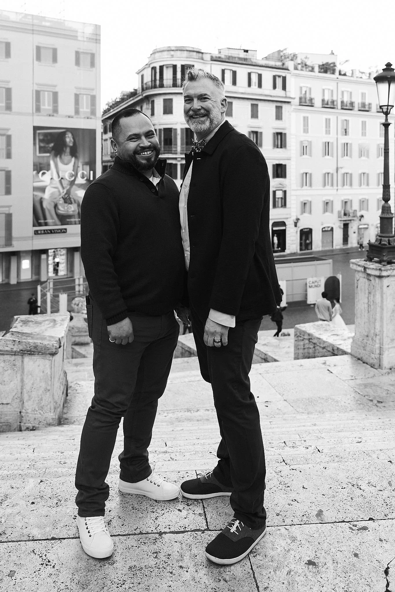 Two men standing outdoors on steps in an urban setting, smiling and posing for a photo. One man has a beard and is wearing a black sweater, dark pants, and white sneakers. The other has light hair and a beard, and is wearing a dark blazer, white shir