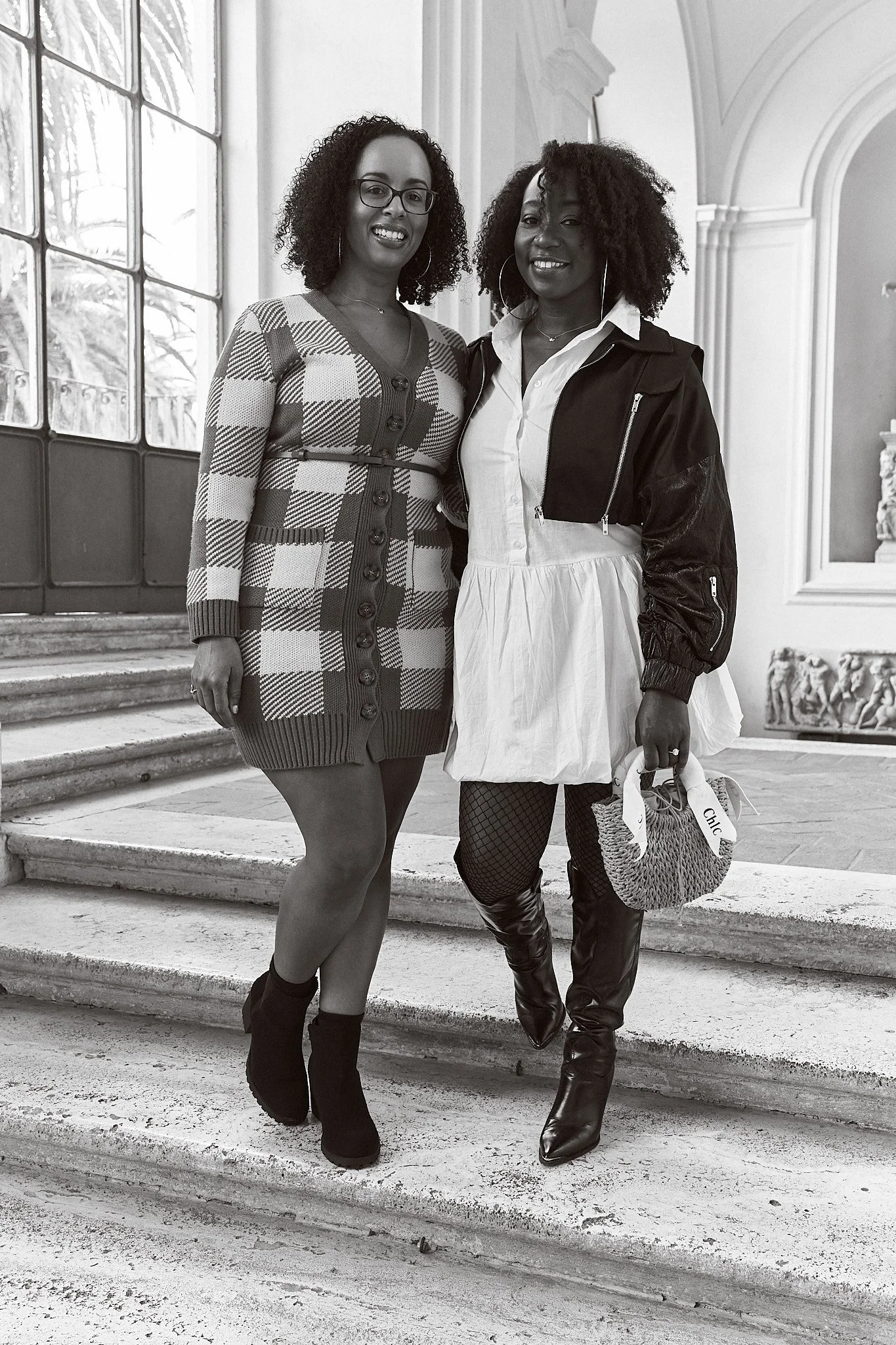 Two women standing on steps inside a large building with arched doorway, large window, and ornate decor in the background, smiling at the camera.
