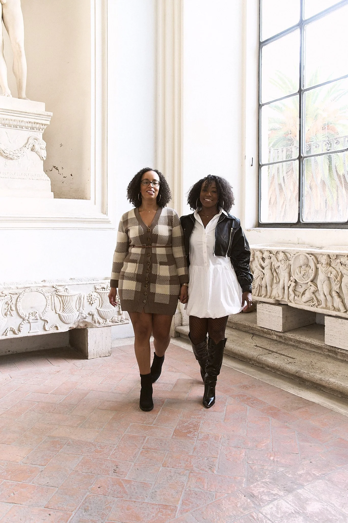Two women walking indoors near a large window and classical sculpture. One is wearing a plaid dress with black boots, the other is wearing a white dress with a black leather jacket and black boots.