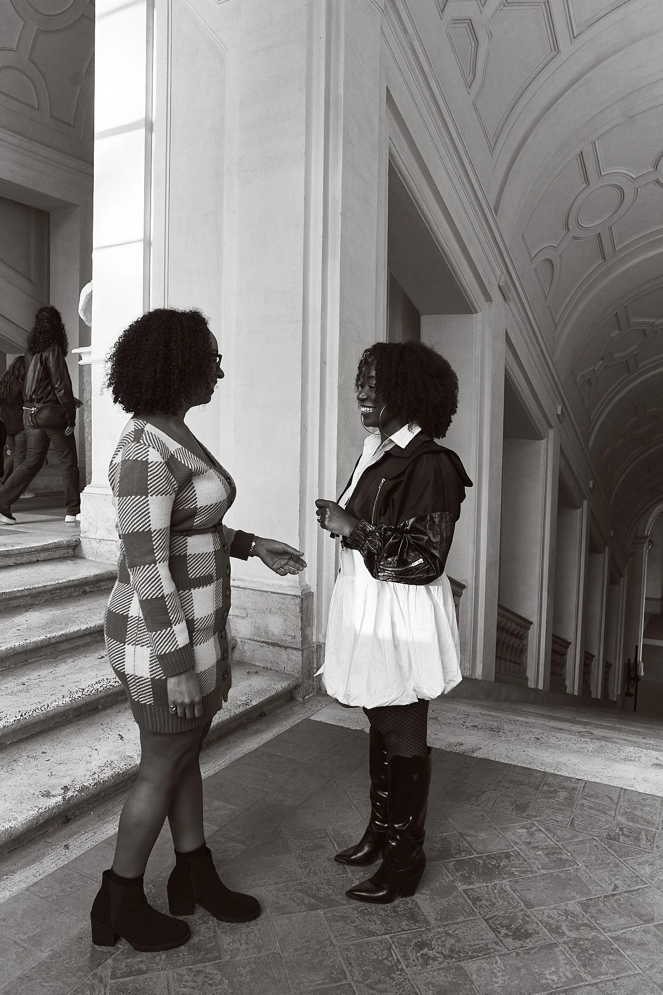 Two women with curly hair and dark skin talking outdoors near stairs, dressed fashionably, wearing boots, one in a checkered blazer, and the other in a fun skirt and leather jacket, with a group of people walking in the background.