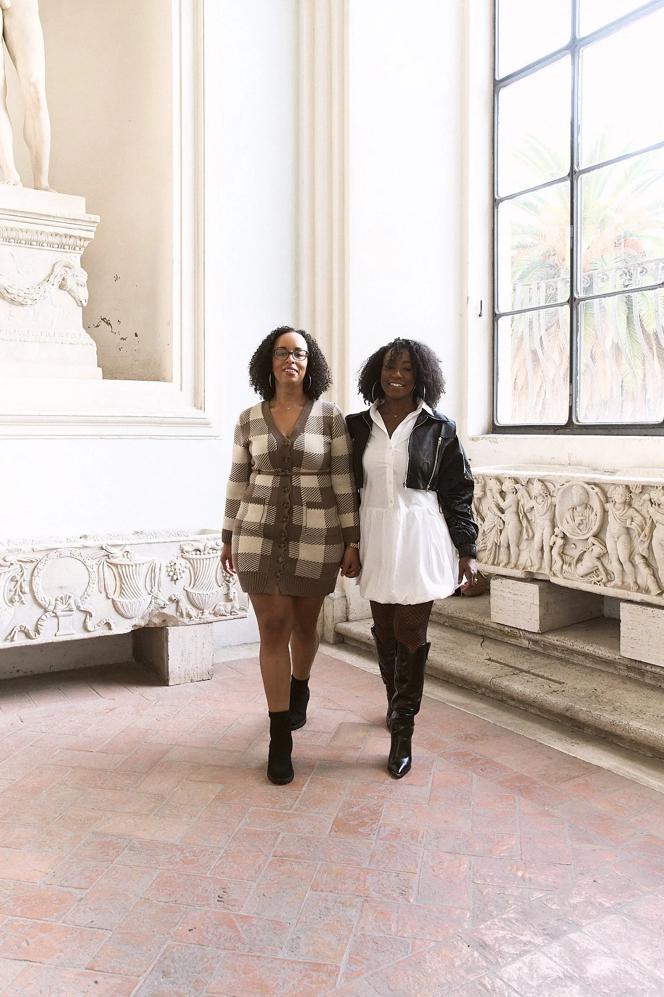 Two women standing side by side in a bright, ornate interior with large window and decorative stone details. They are smiling and dressed in fashion-forward outfits, one in a checked sweater dress and the other in a white dress with a black jacket.