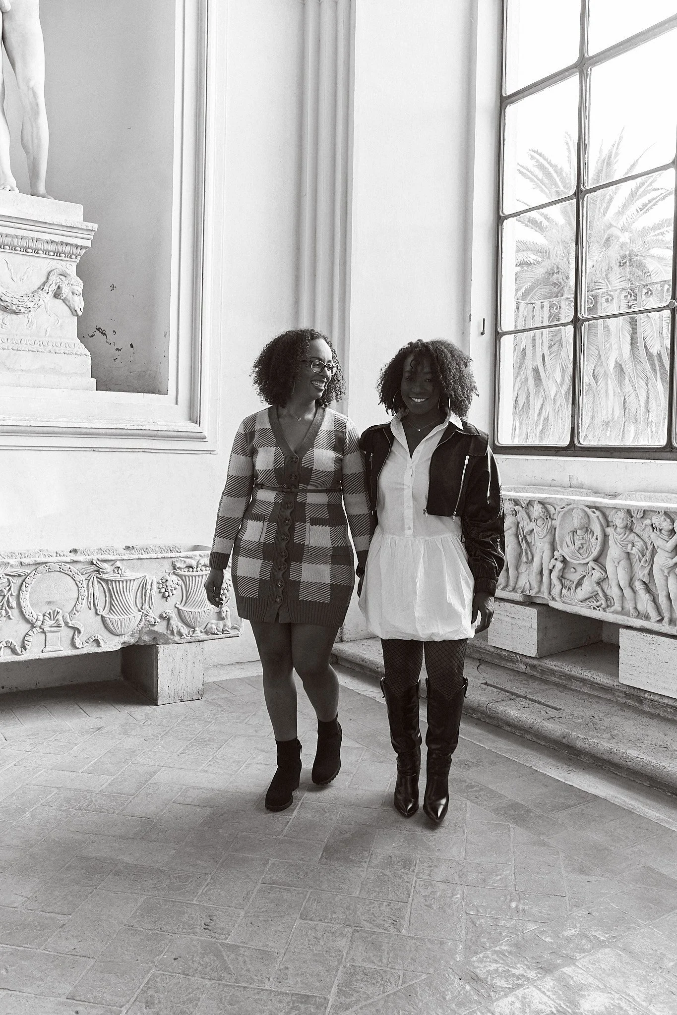 Two women standing inside a historic building with large windows and classical sculptures, smiling at each other.