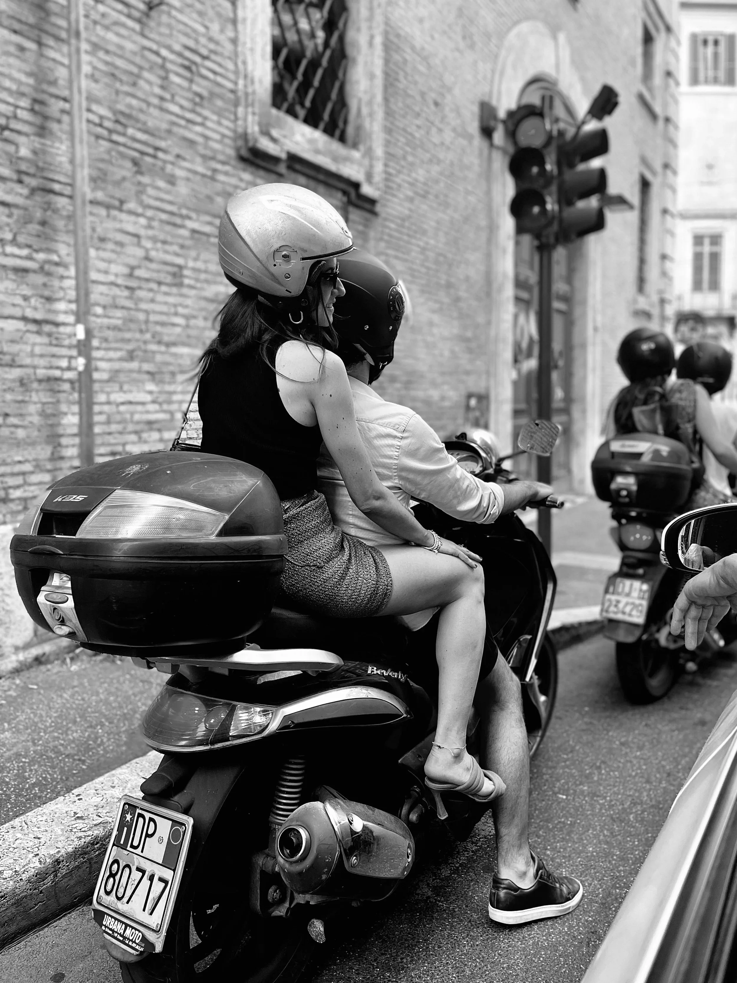 A woman wearing a helmet and sunglasses riding a scooter, with her left hand on the handlebar and foot on the ground, on a city street with brick buildings and traffic lights.