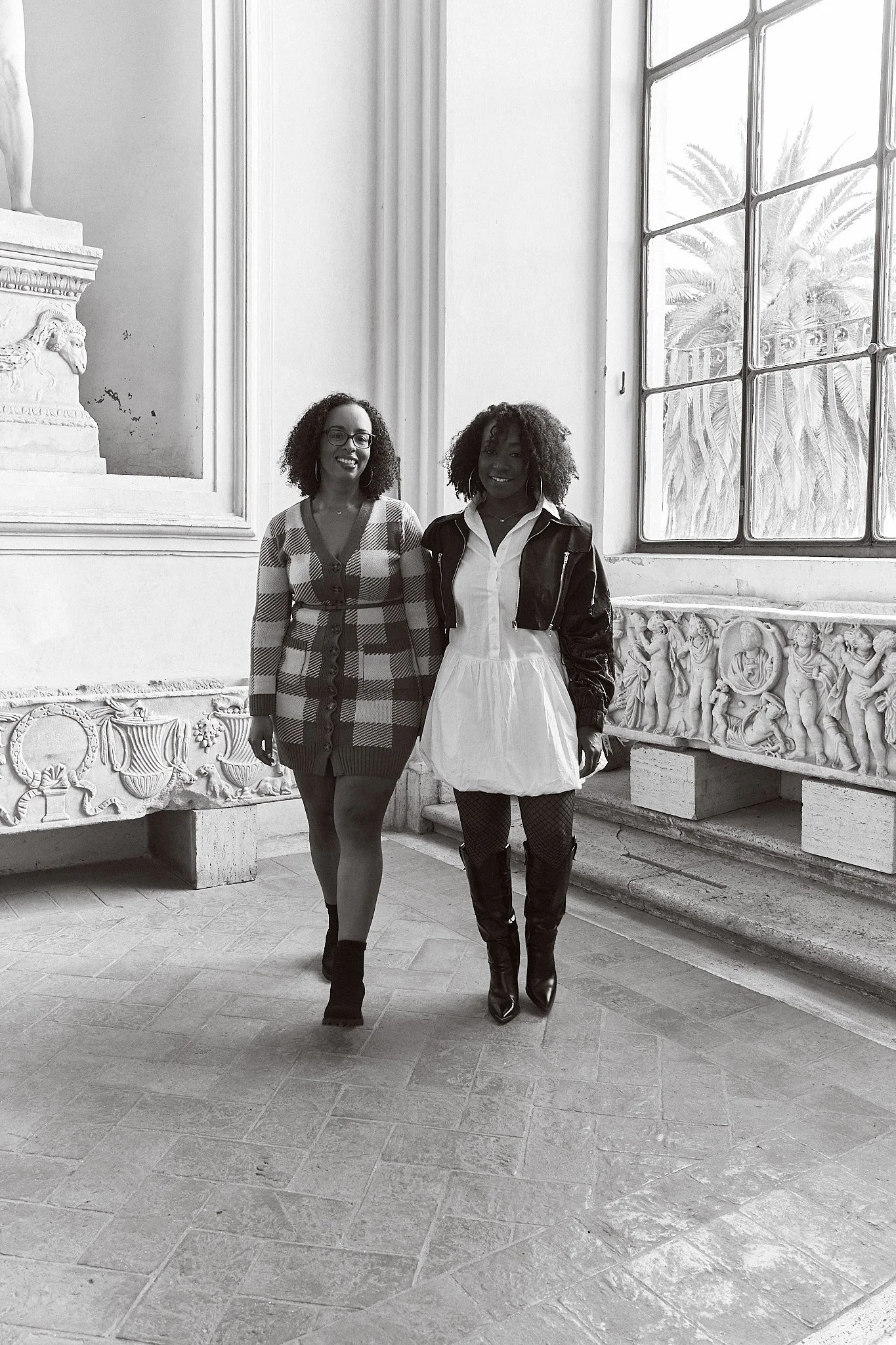 Two women standing inside a historic building with classical sculptures and intricate relief decorations. The woman on the left wears a checkered dress and glasses, and the woman on the right wears a white dress with a leather jacket. They are both s