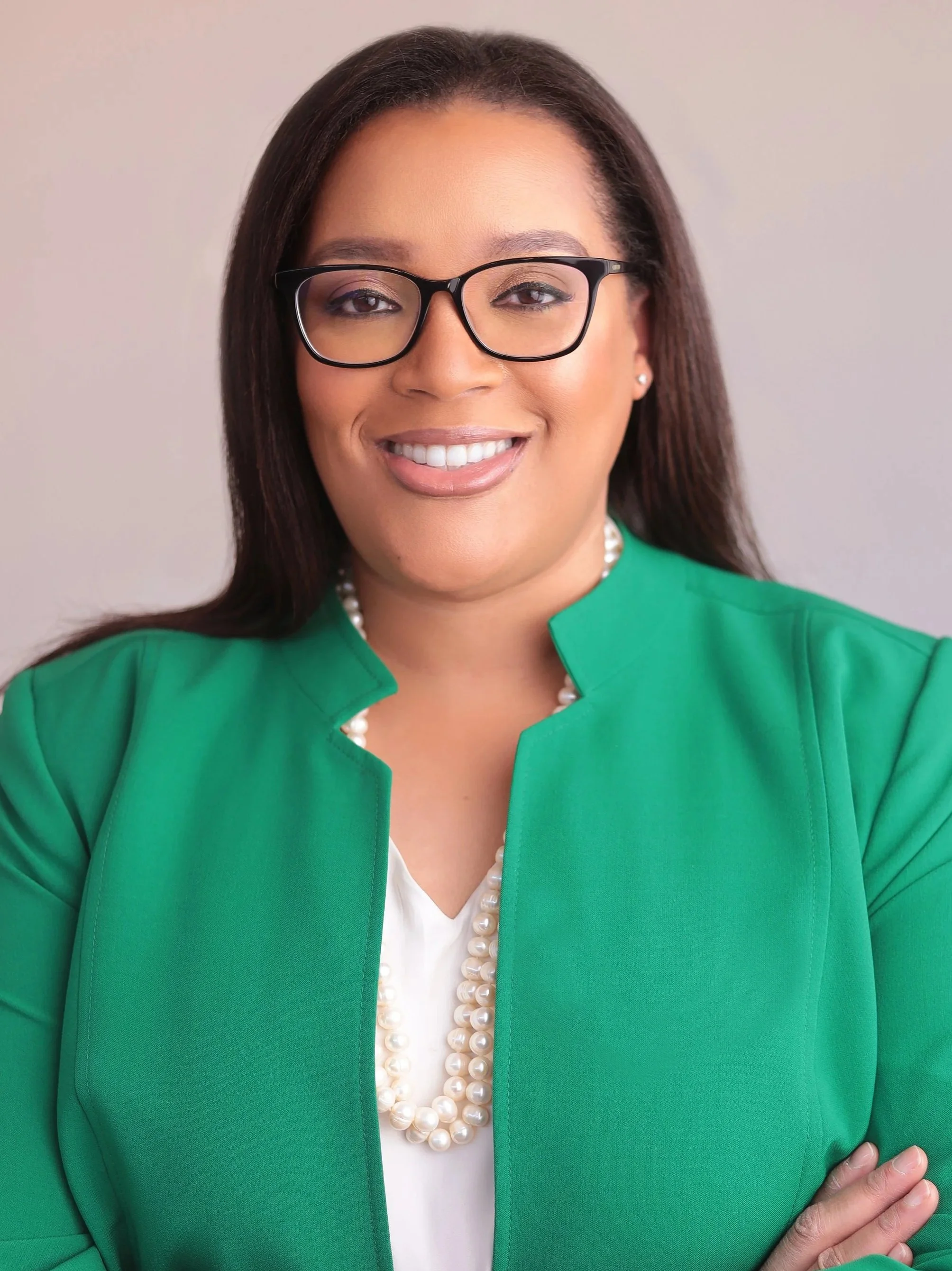 Tabitha Williams, Founder and CEO of Williams Exec Solutions. A woman wearing black glasses, a green blazer, a pearl necklace, and a white top, smiling with her arms crossed against a neutral background.