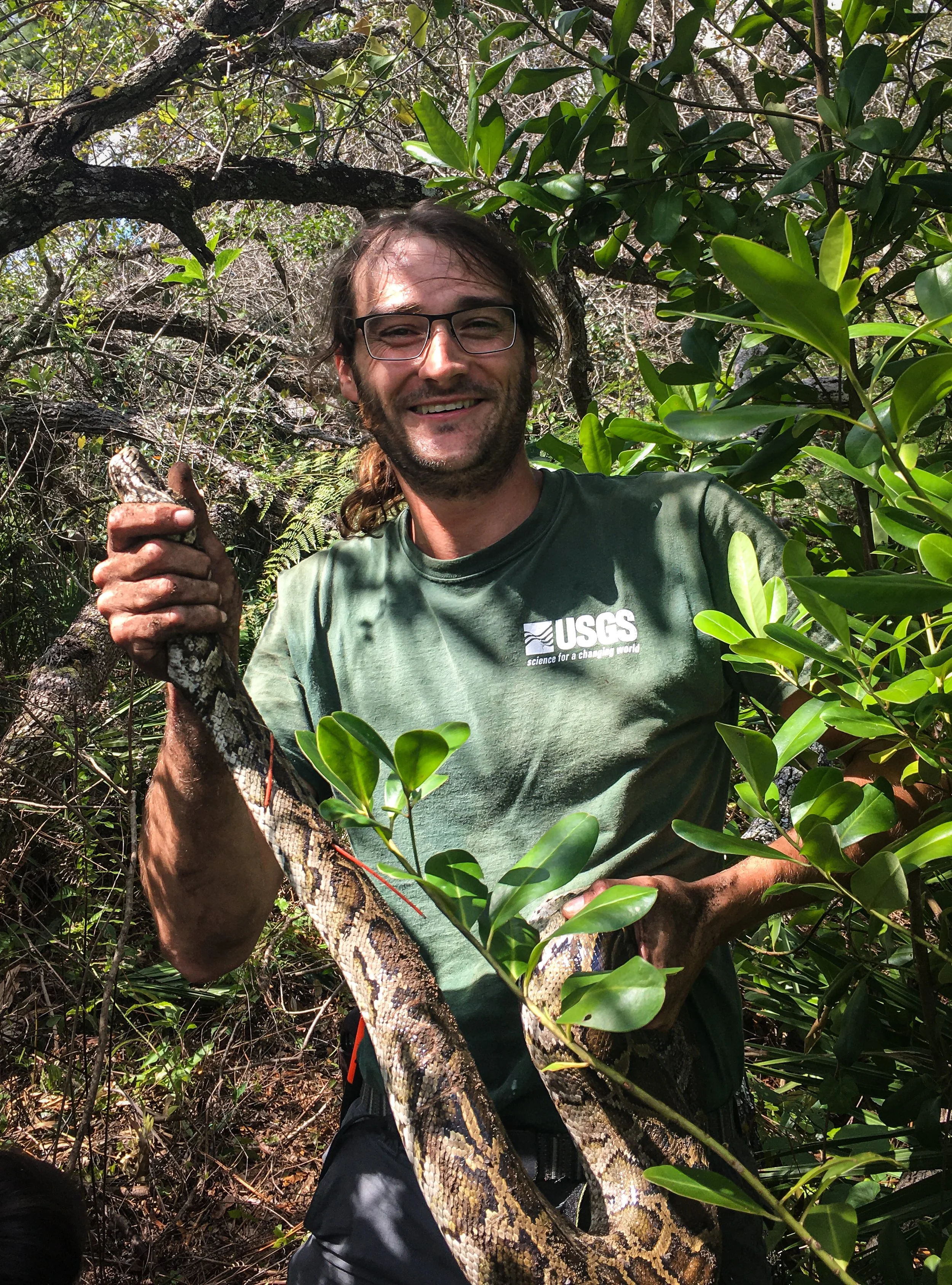 A man with glasses smiling in a green forested area, holding a large python snake.