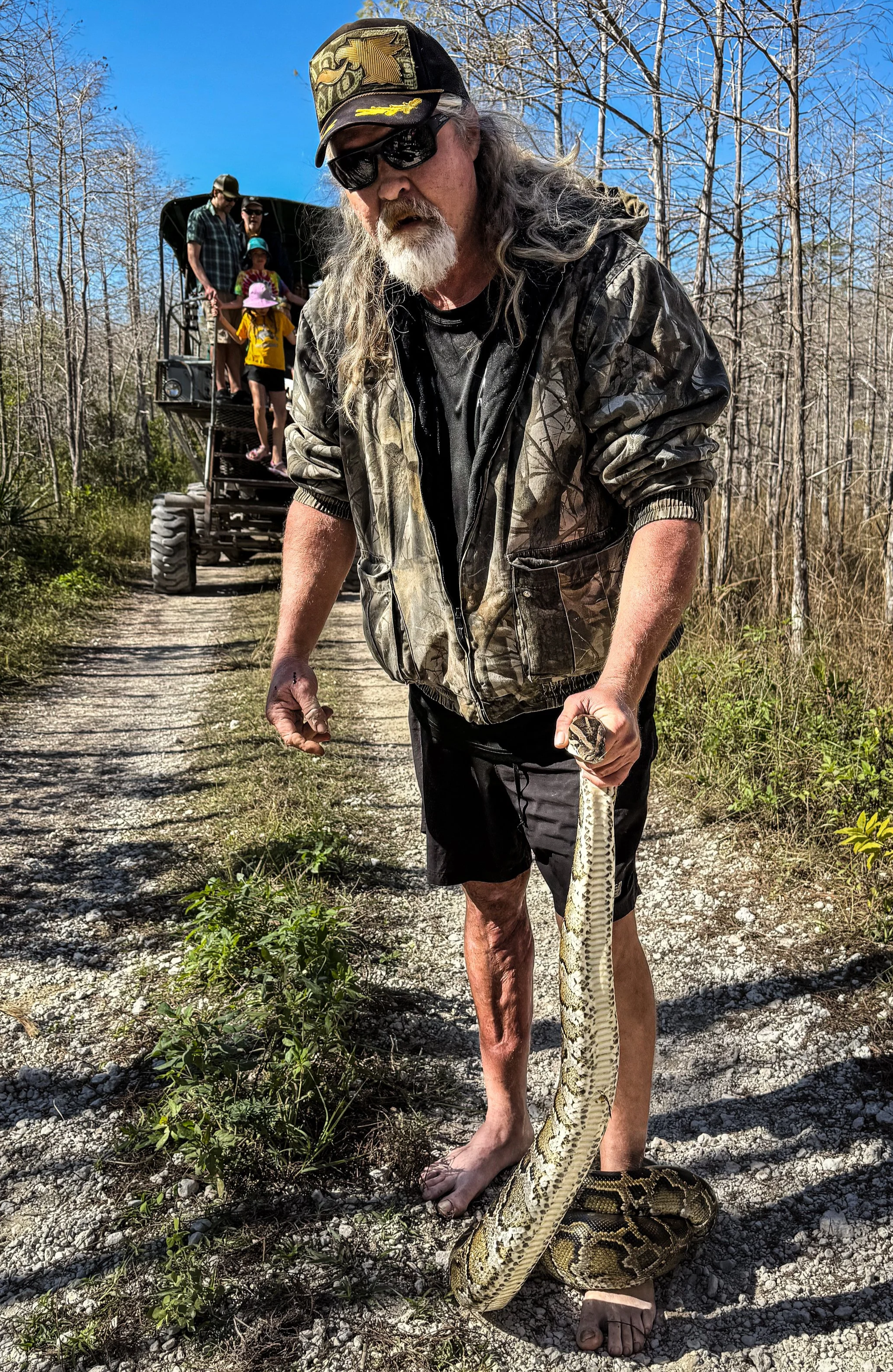 An older man with long gray hair, beard, and sunglasses holding a snake stands barefoot on a gravel trail in a wooded area. In the background, a small group of people on an all-terrain vehicle are visible.