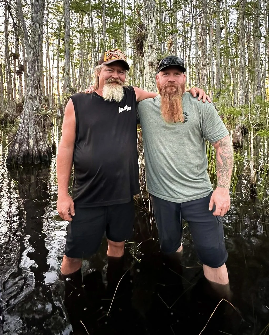 Two men with beards and tattoos standing in a swampy wooded area with water up to their knees, smiling and posing for a photo.