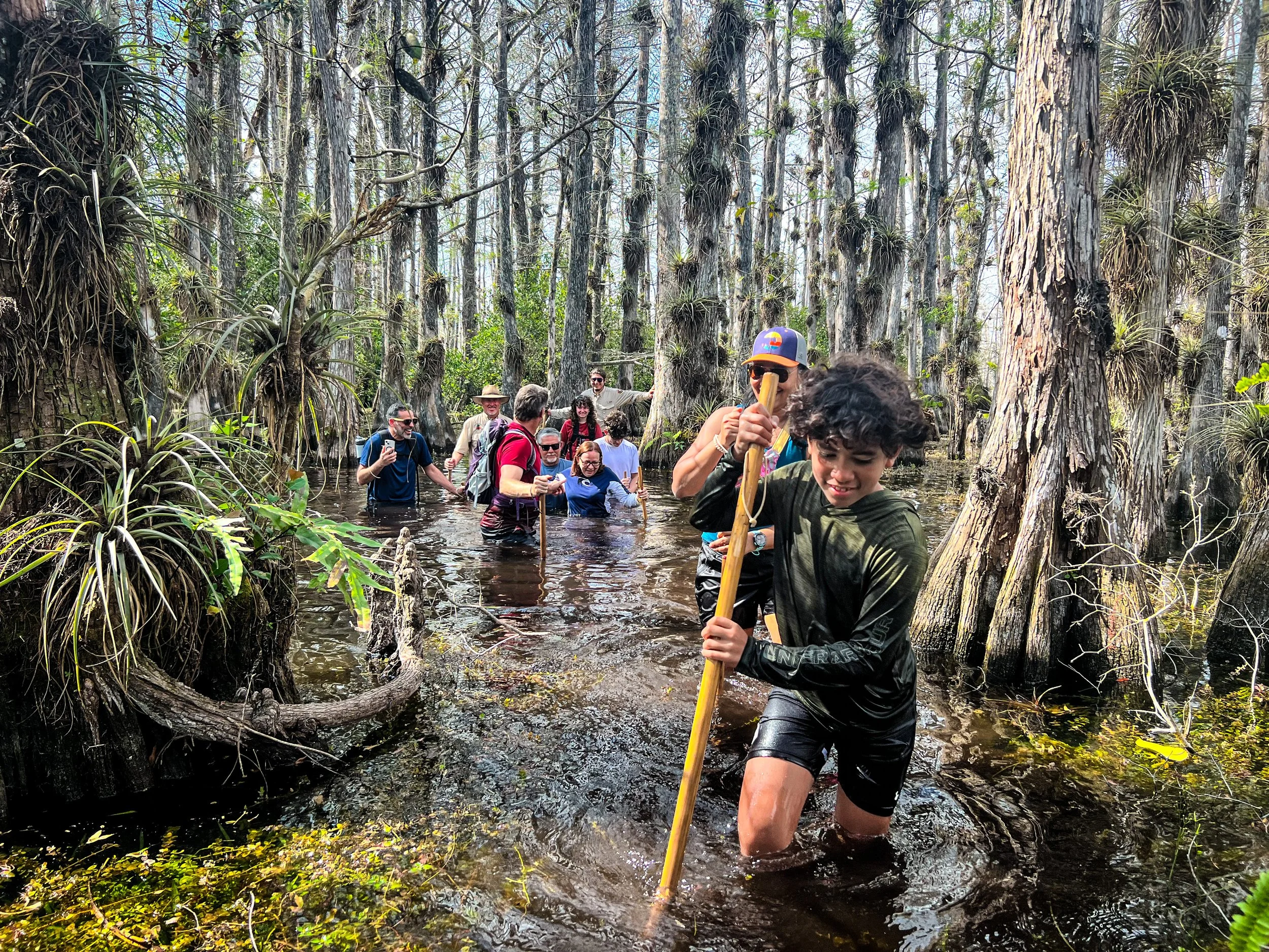 Group of people hiking through a swampy forest, wading through water and surrounded by trees and wetlands.
