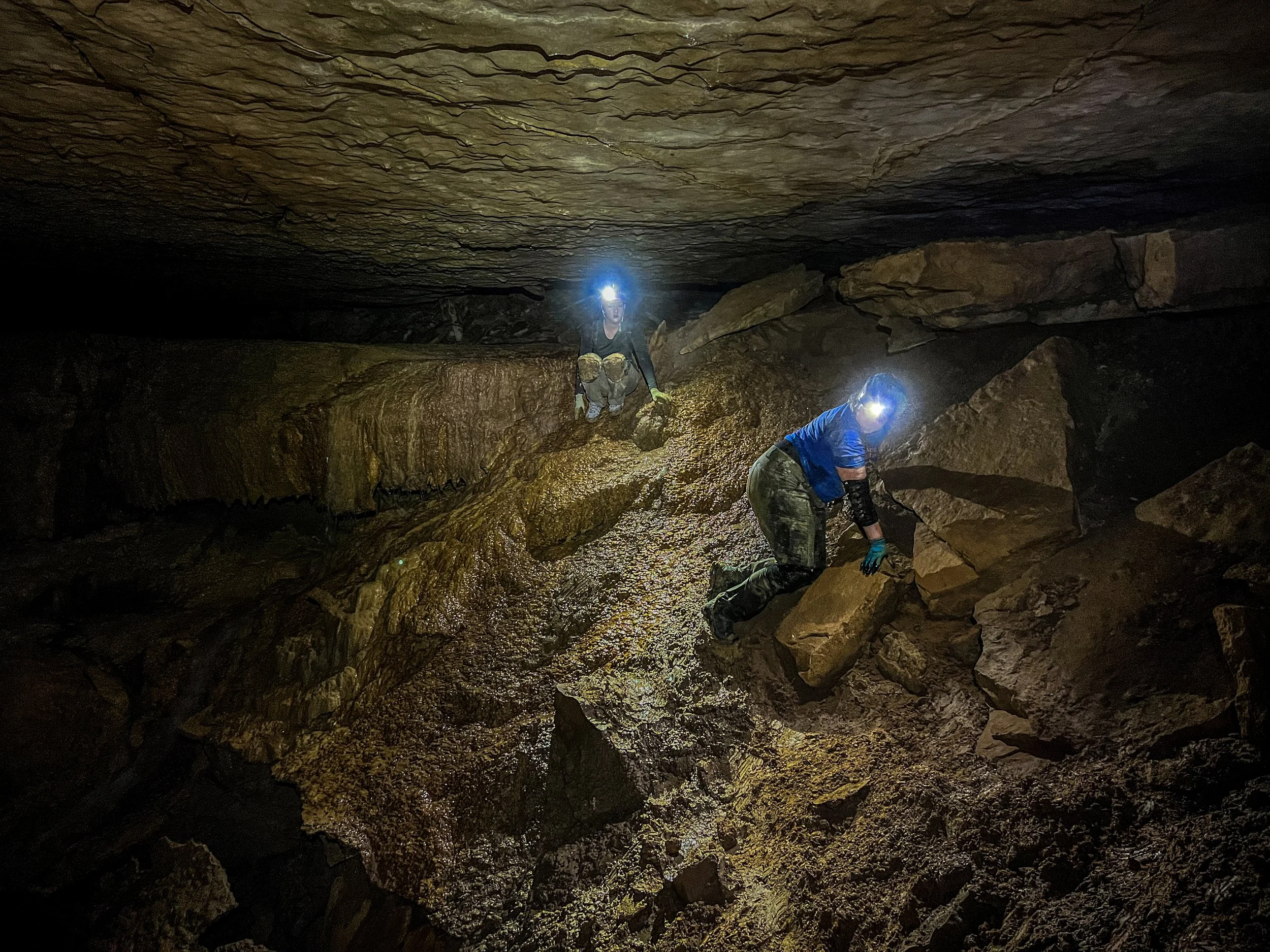 Two cavers wearing helmets with lights explore a dark, rocky underground cave. One is climbing over rocks, and the other is crouching on a sloped surface, both surrounded by large boulders and uneven terrain.