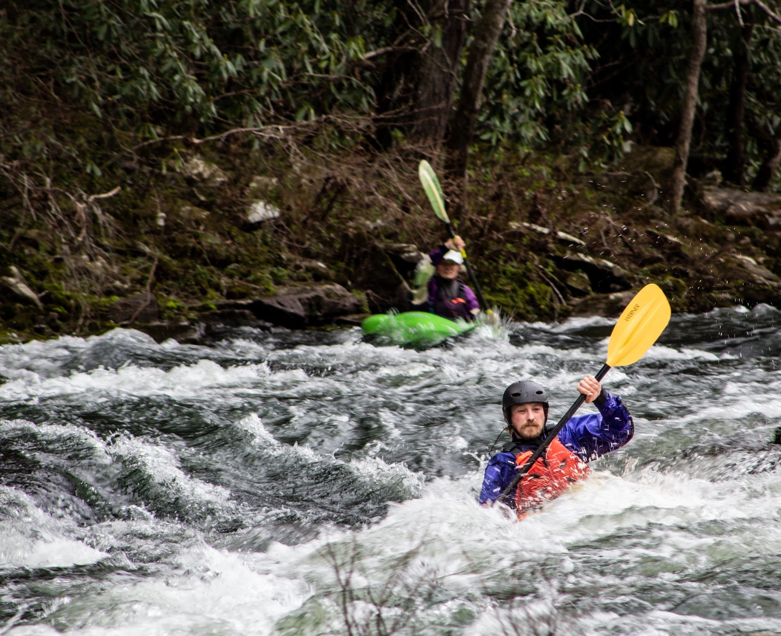 Two kayakers paddling through a rocky river with forested shoreline in the background.
