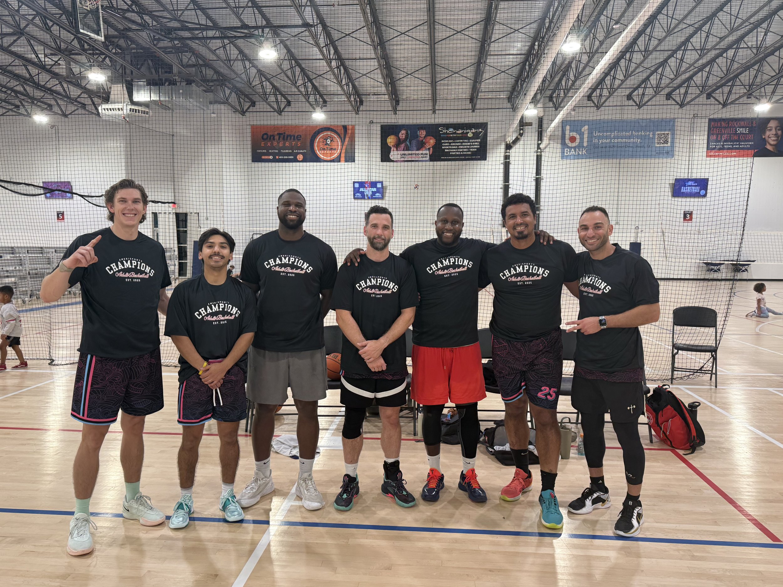 Seven men posing together in an indoor basketball court, wearing matching 'Champions' t-shirts, with some making gestures and smiling, with children playing in the background.