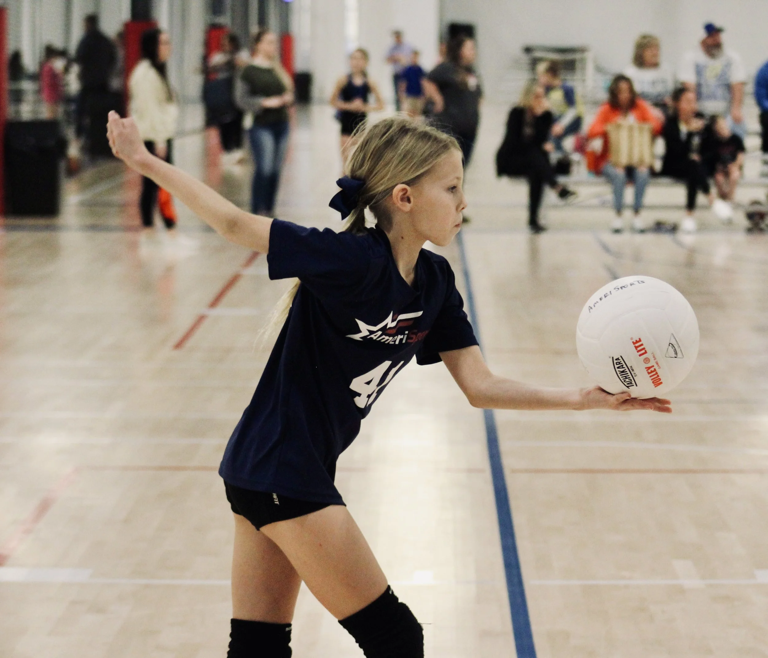 Young girl in a navy sports uniform with a bow in her hair playing volleyball at an indoor gymnasium, with spectators in the background.